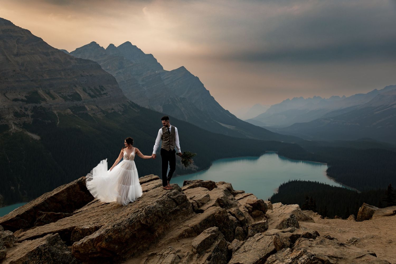 Couple in wedding attire stands on rocky mountain peak overlooking misty alpine lake and mountain ranges at sunset.