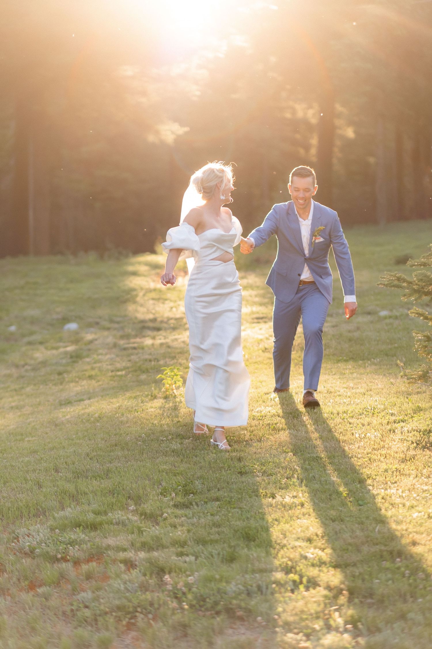 Bride and groom holding hands running through sunlit forest meadow in wedding attire at golden hour.