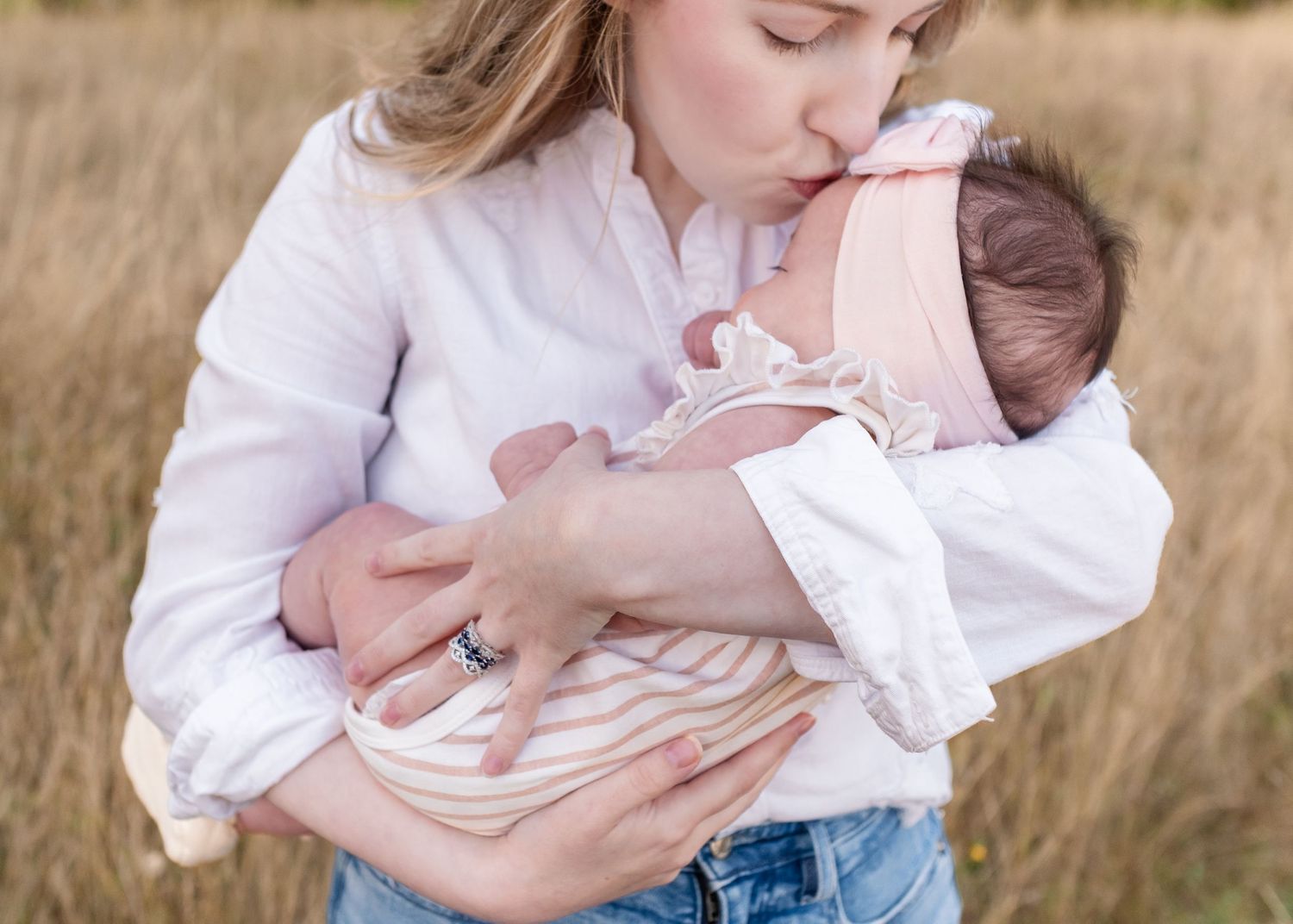 Mother in white blouse holds and tenderly embraces baby in golden sunlit field at sunset.