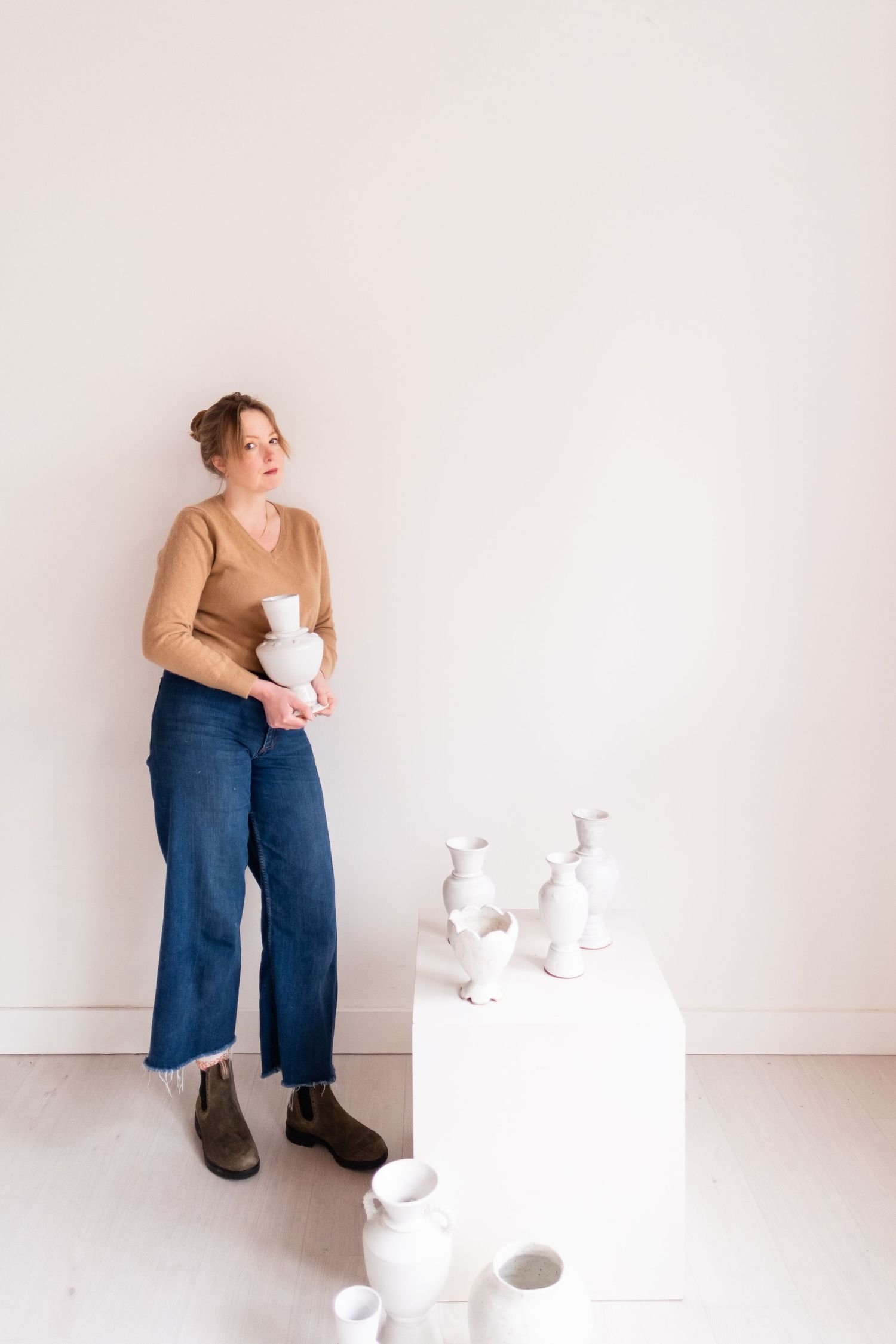 Figure in tan sweater and denim stands beside white pedestal displaying ceramic pieces in bright interior space.