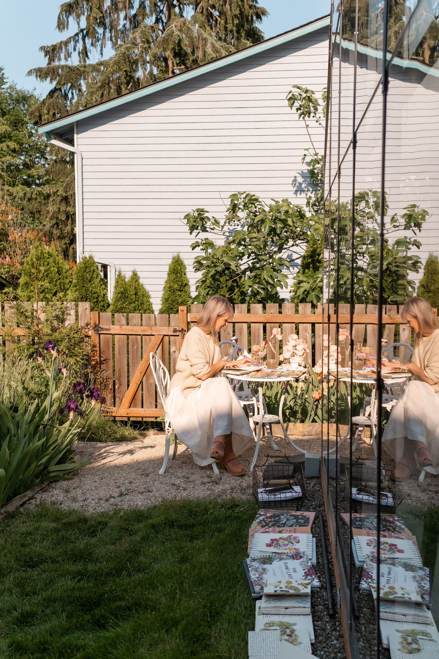 Woman in beige dress painting at a white garden table surrounded by lush greenery and flowers.