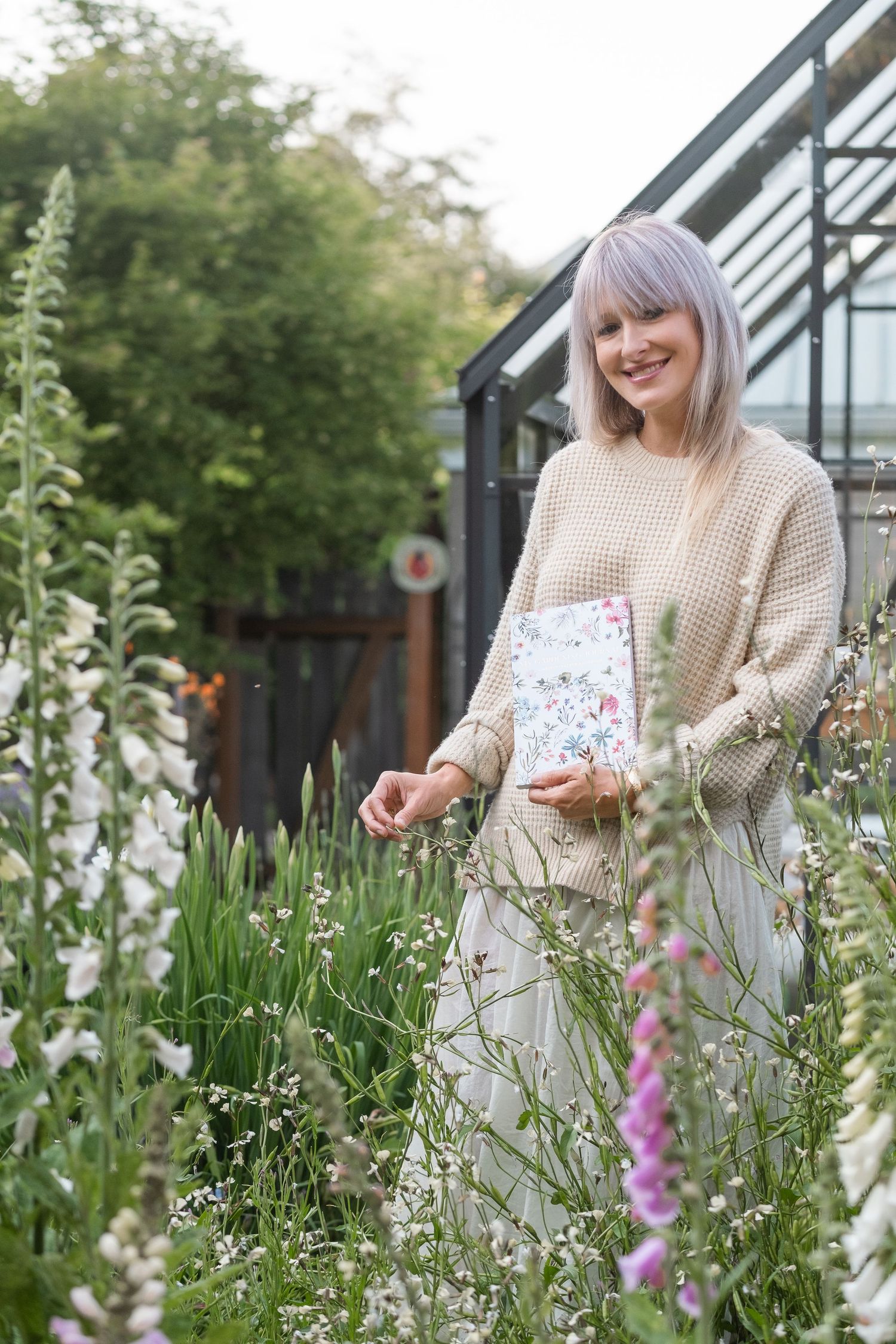 Happy woman in beige sweater holding a floral journal while standing among tall garden wildflowers.