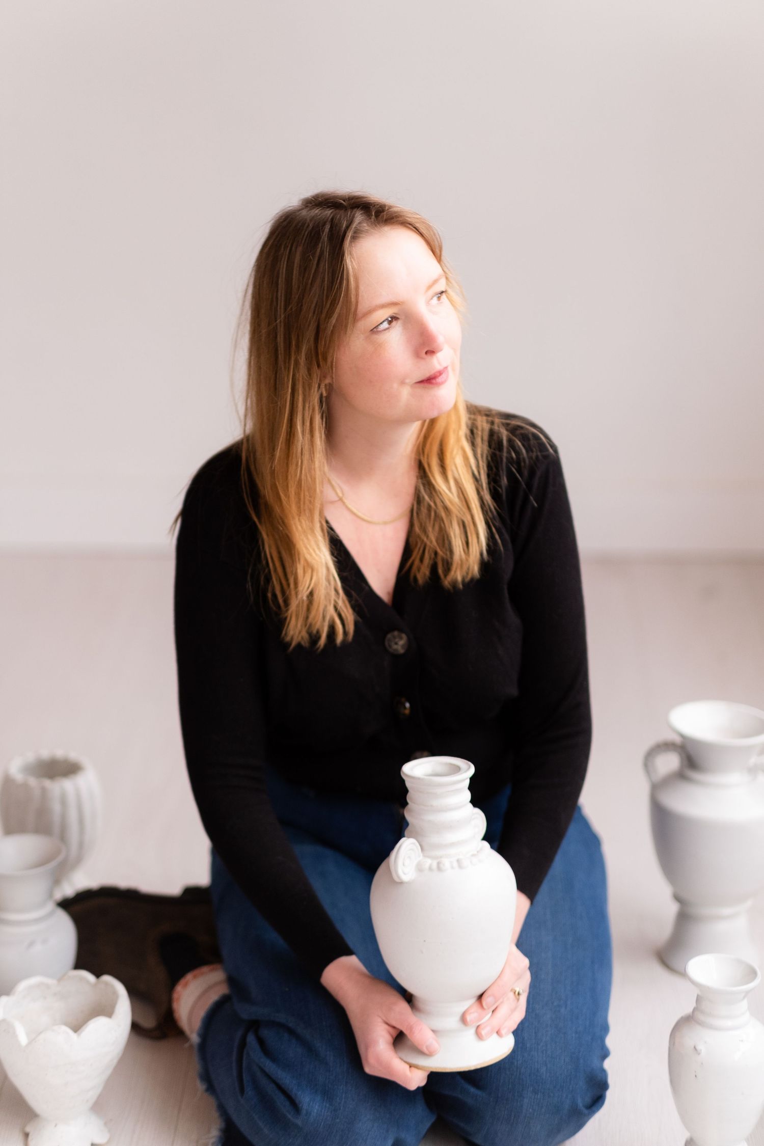 Artist in black cardigan examines white ceramic vase while surrounded by pottery pieces in minimalist studio setting.