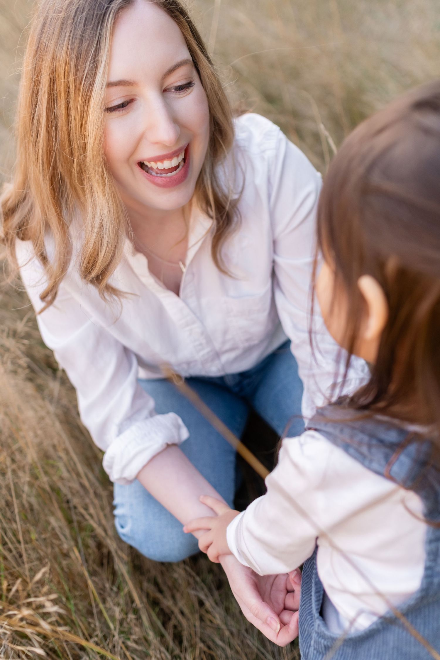 Two people in casual attire share a joyful moment while sitting in tall grass on a sunny day.