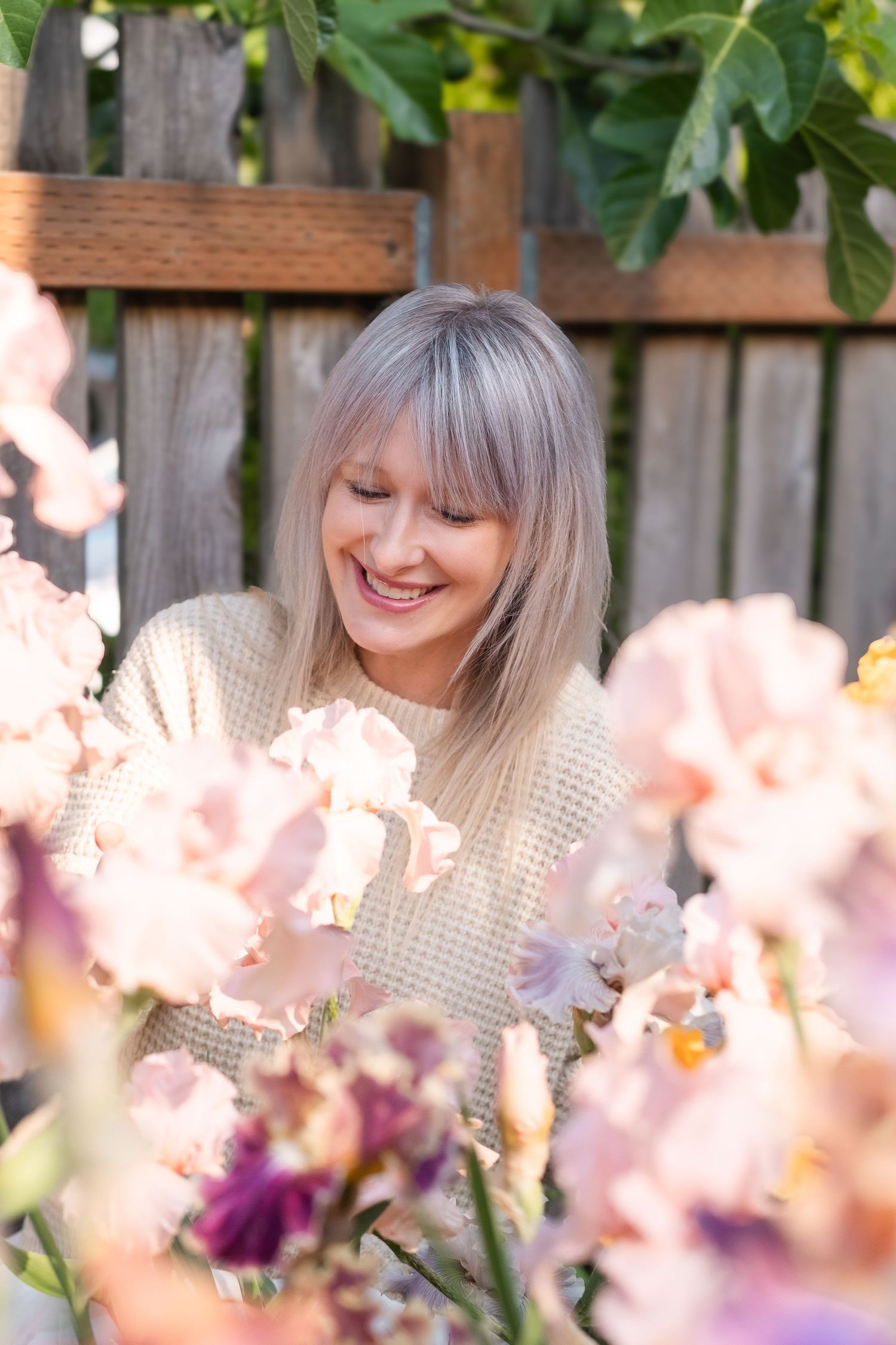 Smiling blonde woman surrounded by blooming pink and purple iris flowers in a garden.