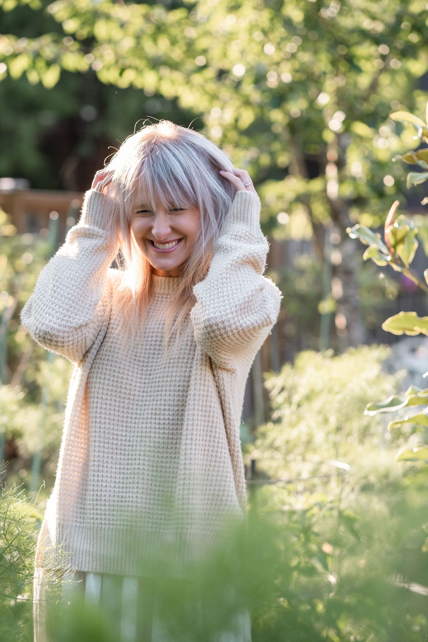 Joyful blonde woman in beige waffle knit sweater laughing in a sunny green garden.