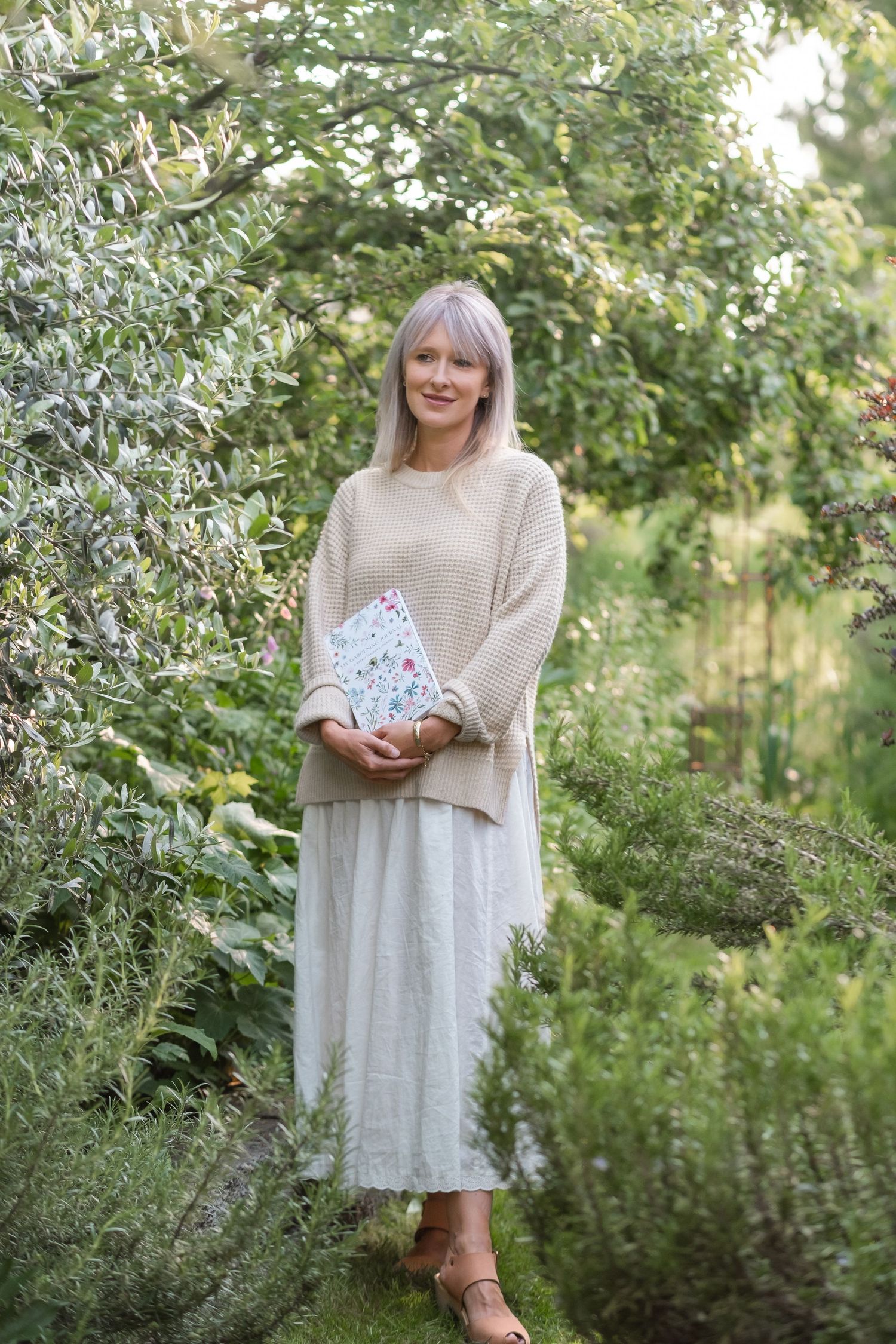 Smiling blonde woman holding floral notebook standing among lush green garden plants.