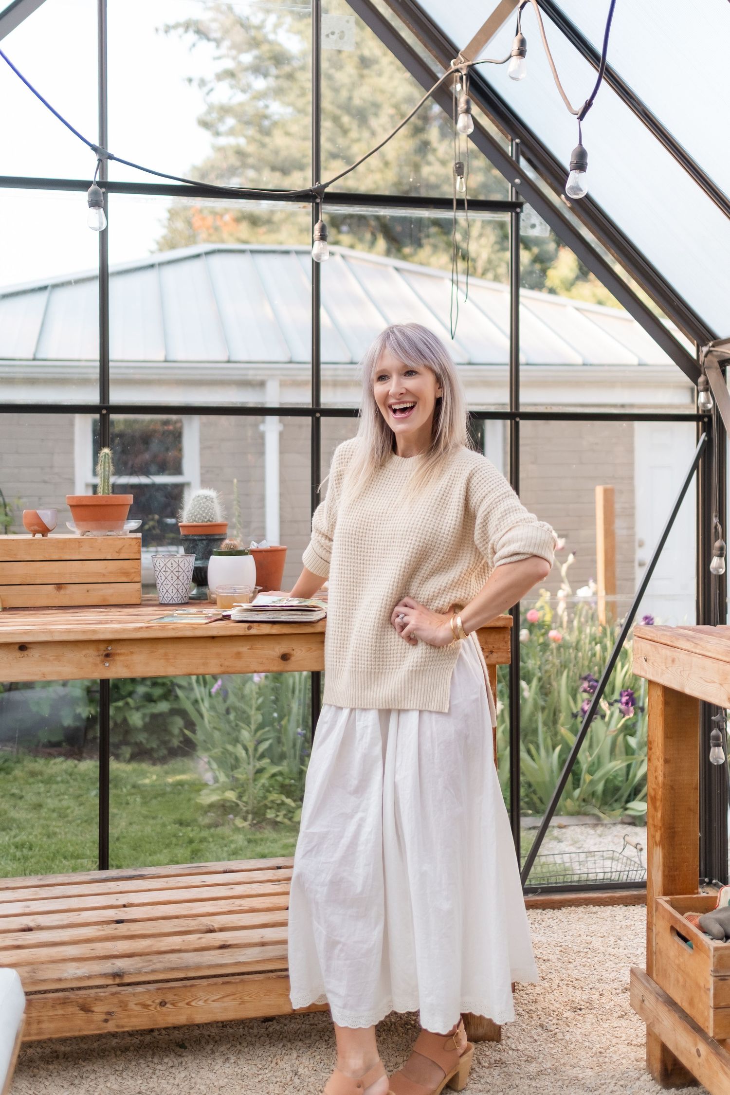 Laughing blonde woman in beige sweater and white skirt standing inside a glass greenhouse.