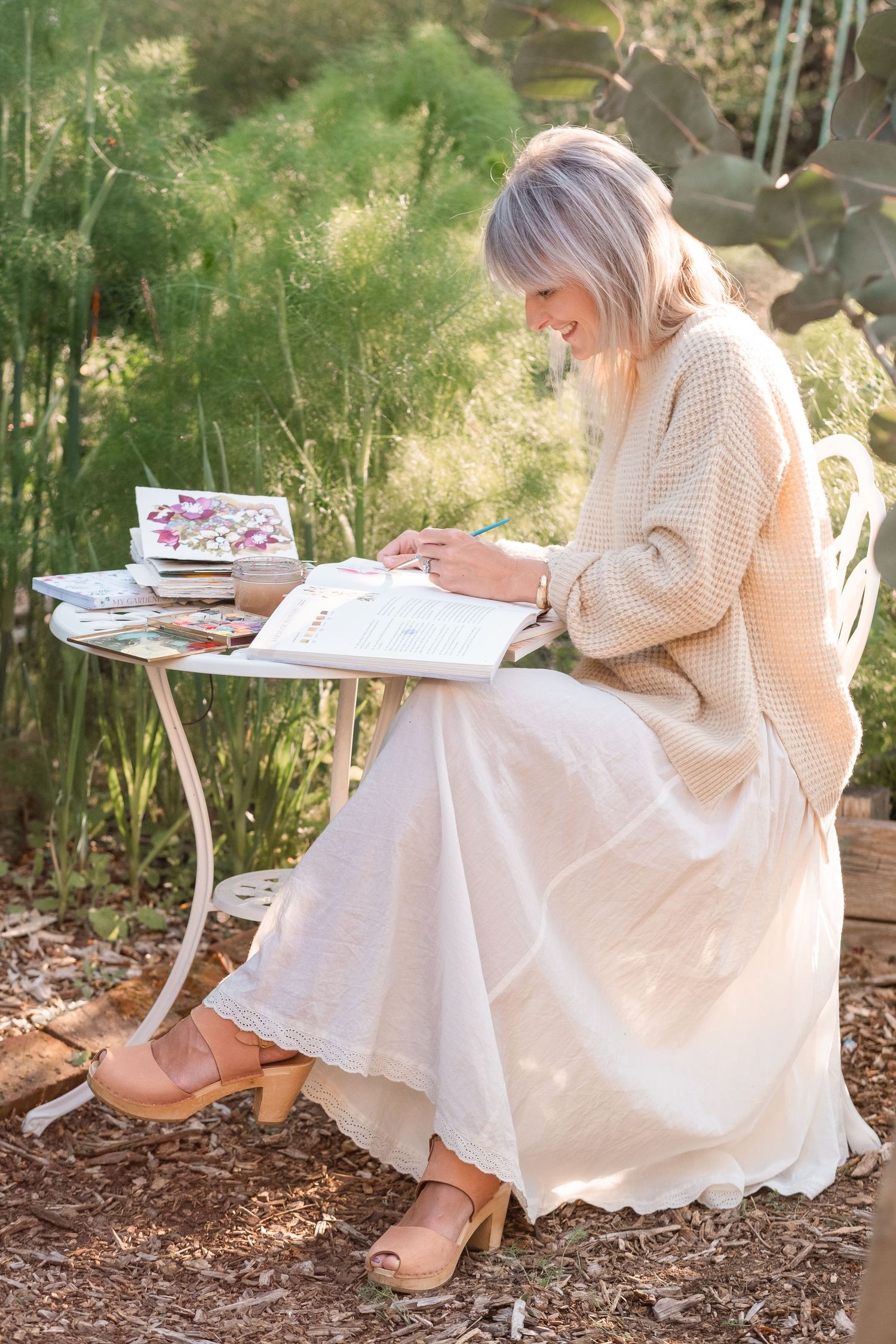 Woman in cream knit sweater and linen skirt sitting at a garden table writing in an open journal.