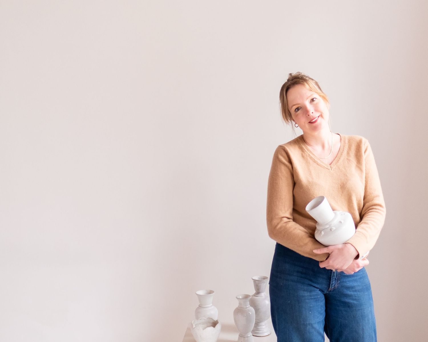 A person in a tan sweater and jeans holds a white ceramic vase against a light pink background.