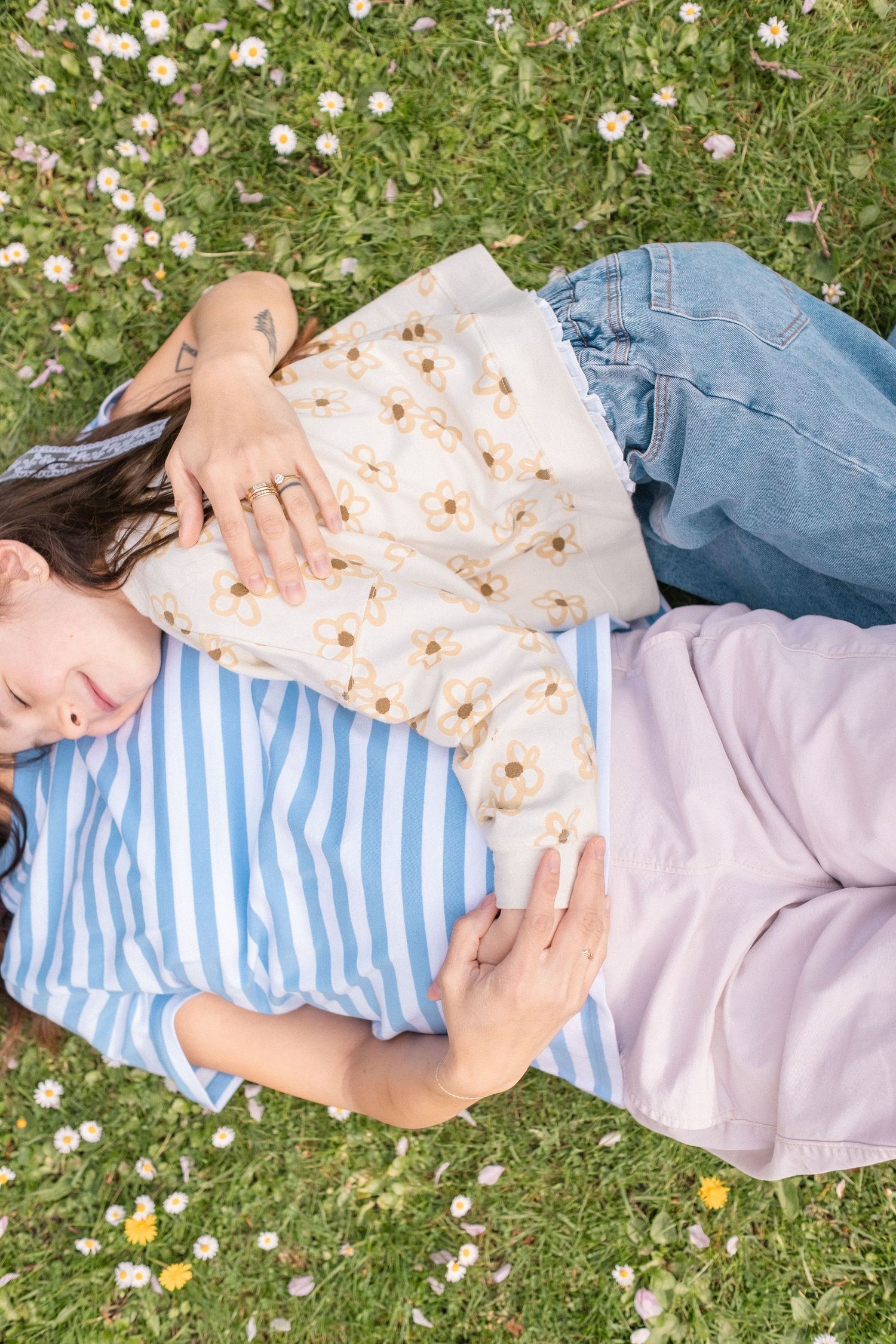 Someone lying on grass scattered with daisies wearing a striped shirt, floral top and jeans on a sunny day.
