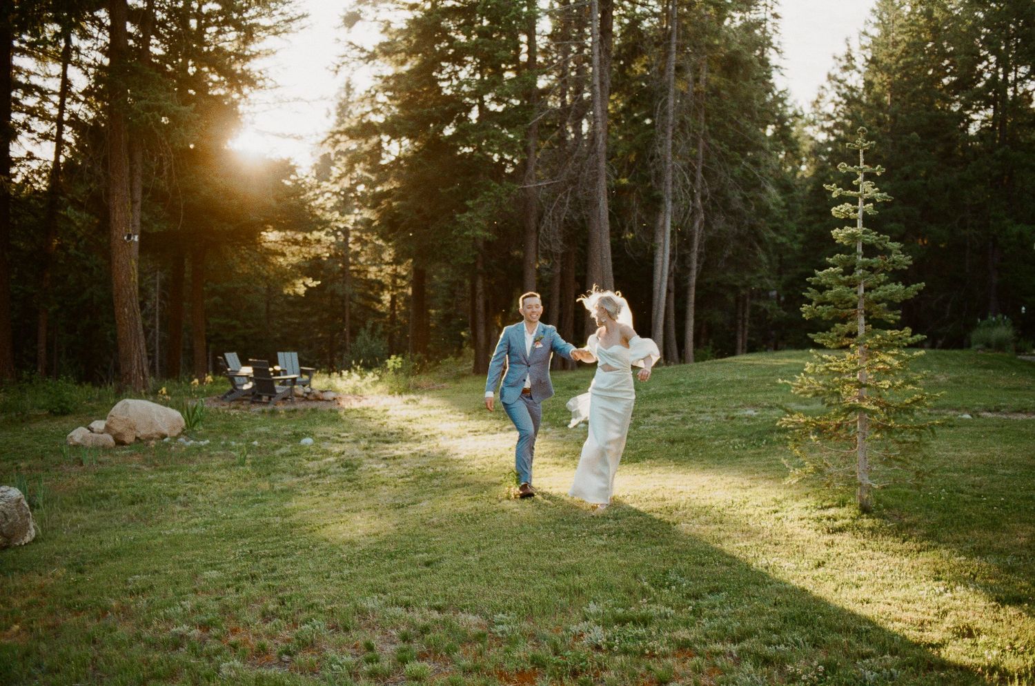 Couple dancing joyfully on green lawn surrounded by tall pine trees during golden hour sunset at outdoor wedding.