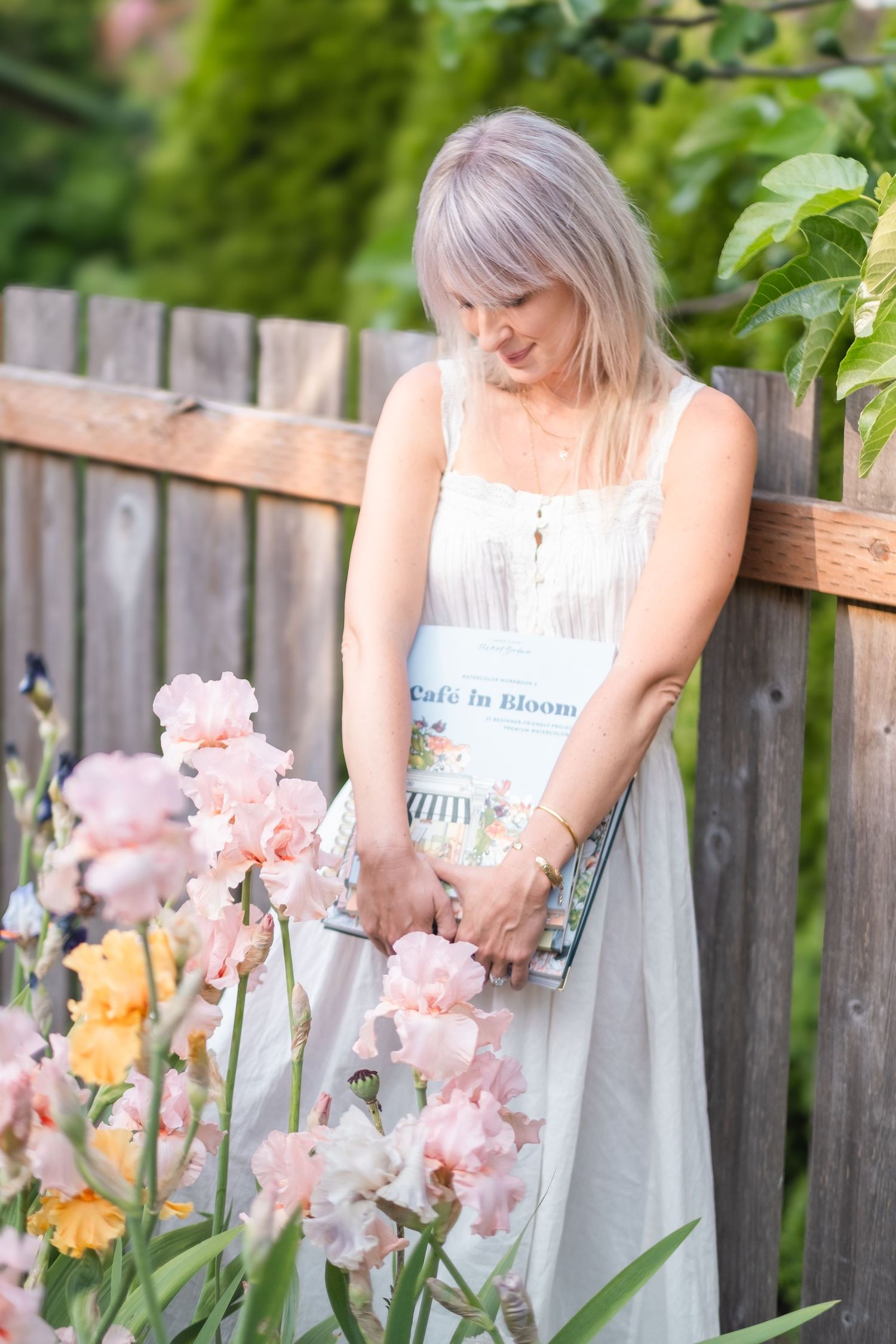 Woman in white dress holding Cafe in Bloom book among pink iris flowers by wooden fence.