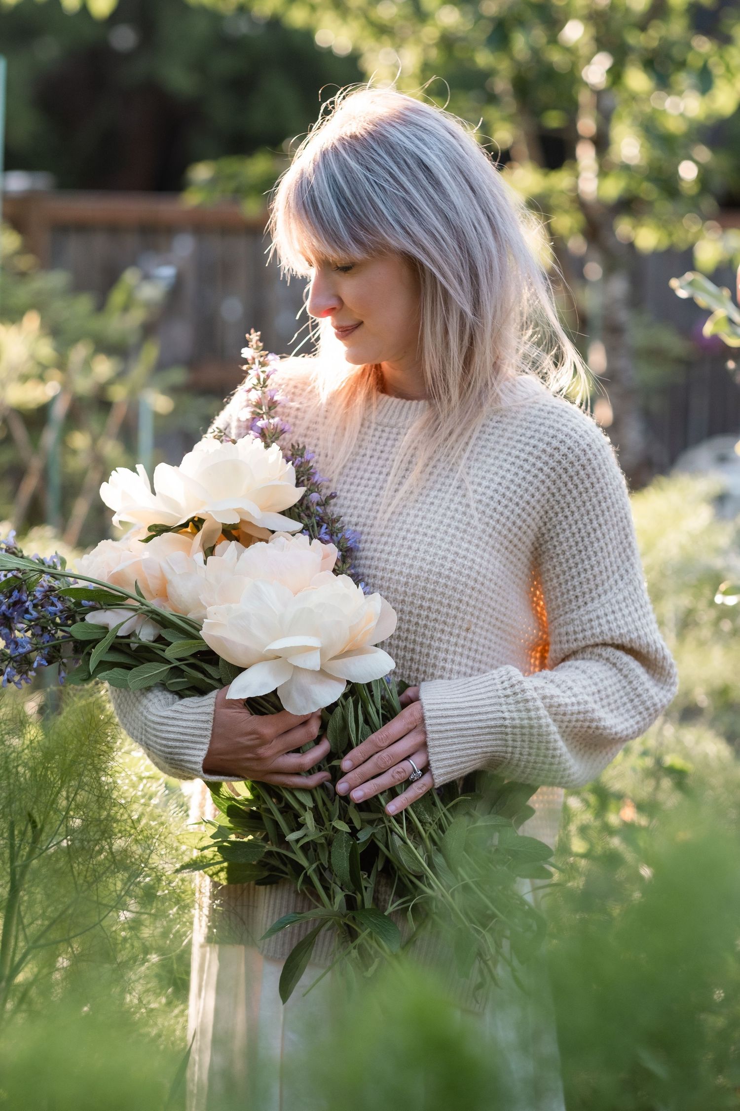 Woman with blonde hair holding a bouquet of white peonies and purple flowers in a sunlit garden.