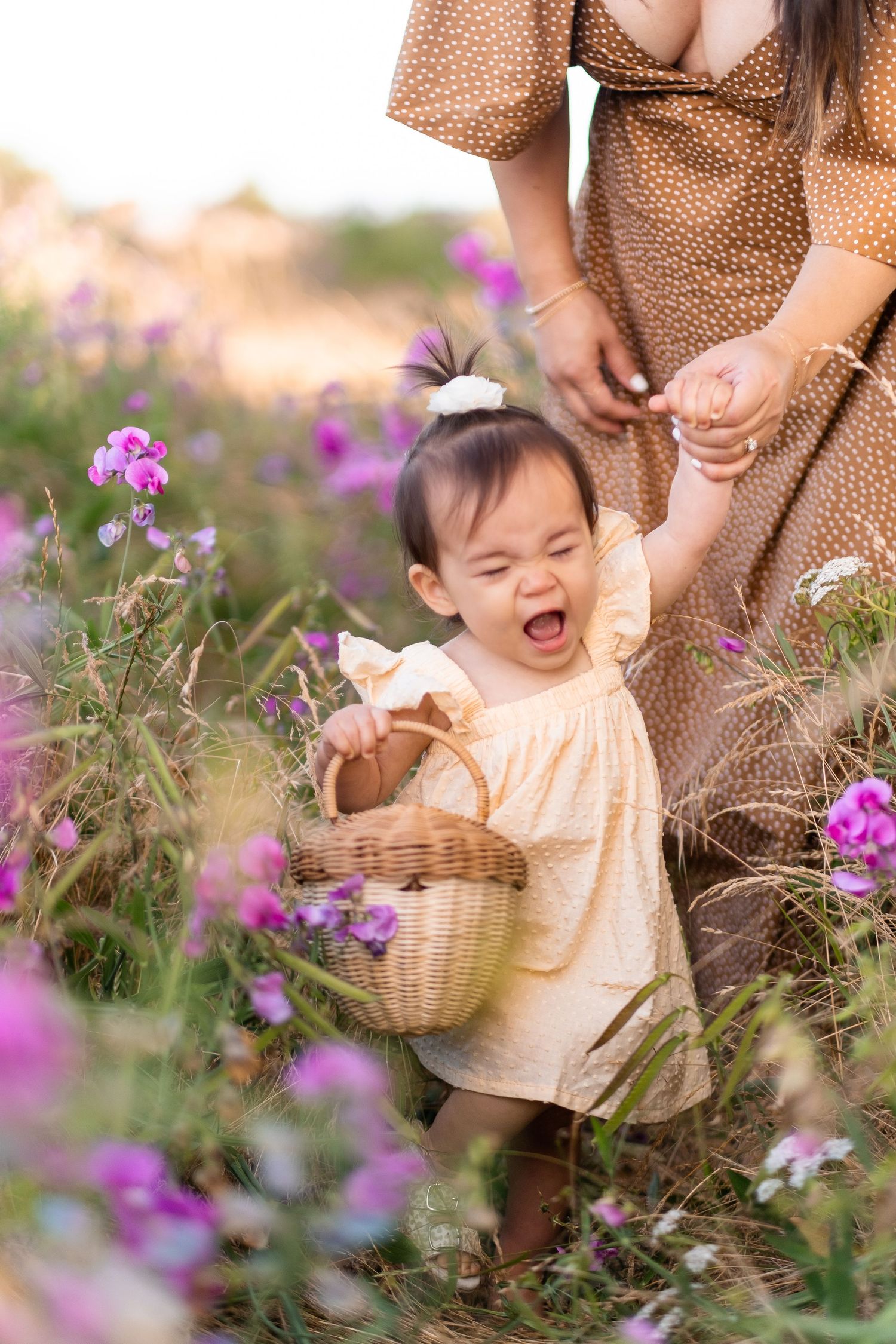 A small baby wearing cream dress holding basket yawns while standing in field of purple wildflowers at sunset.