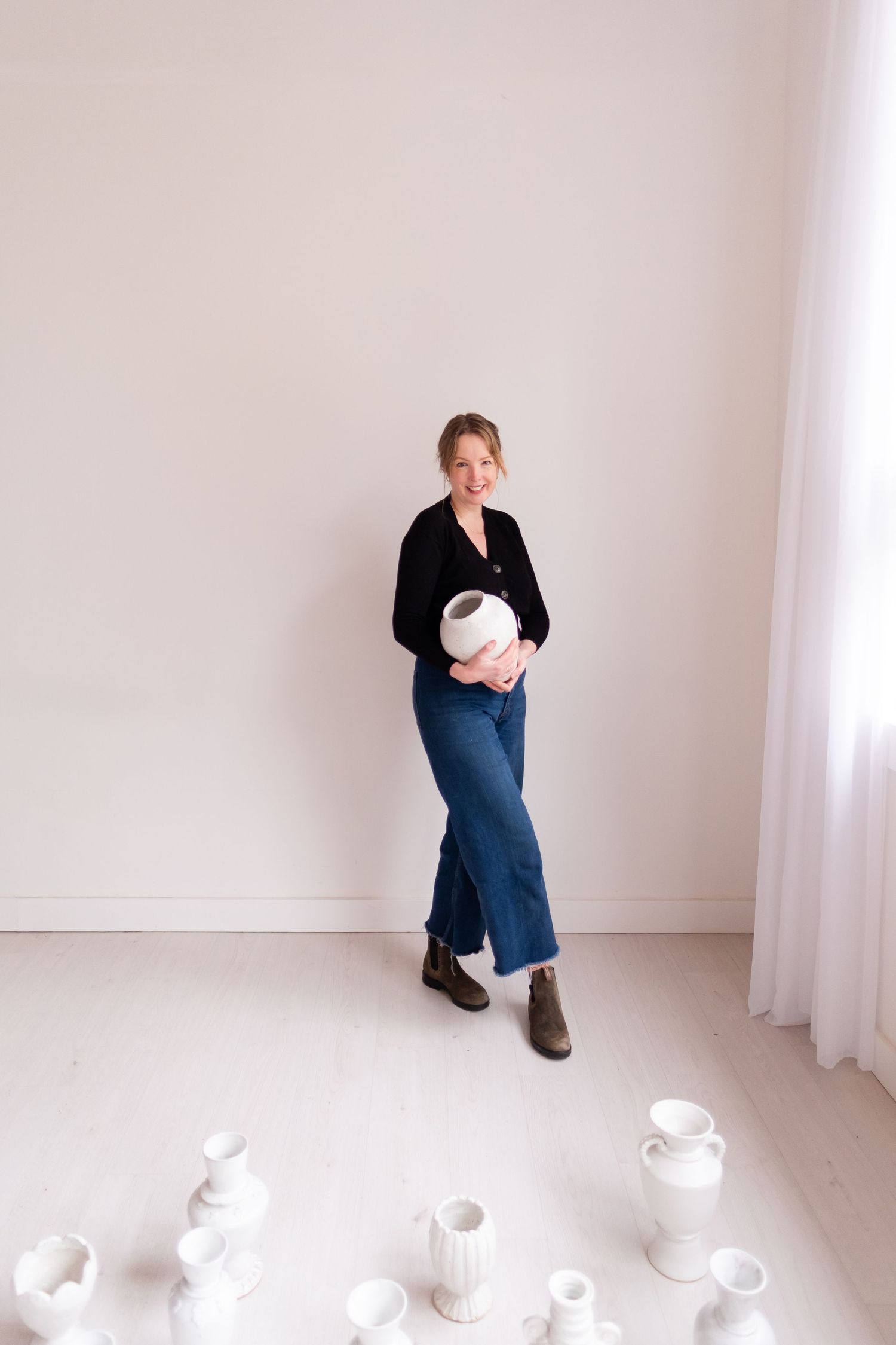 Individual holding white pottery stands in bright minimalist room with ceramic pieces displayed on floor.
