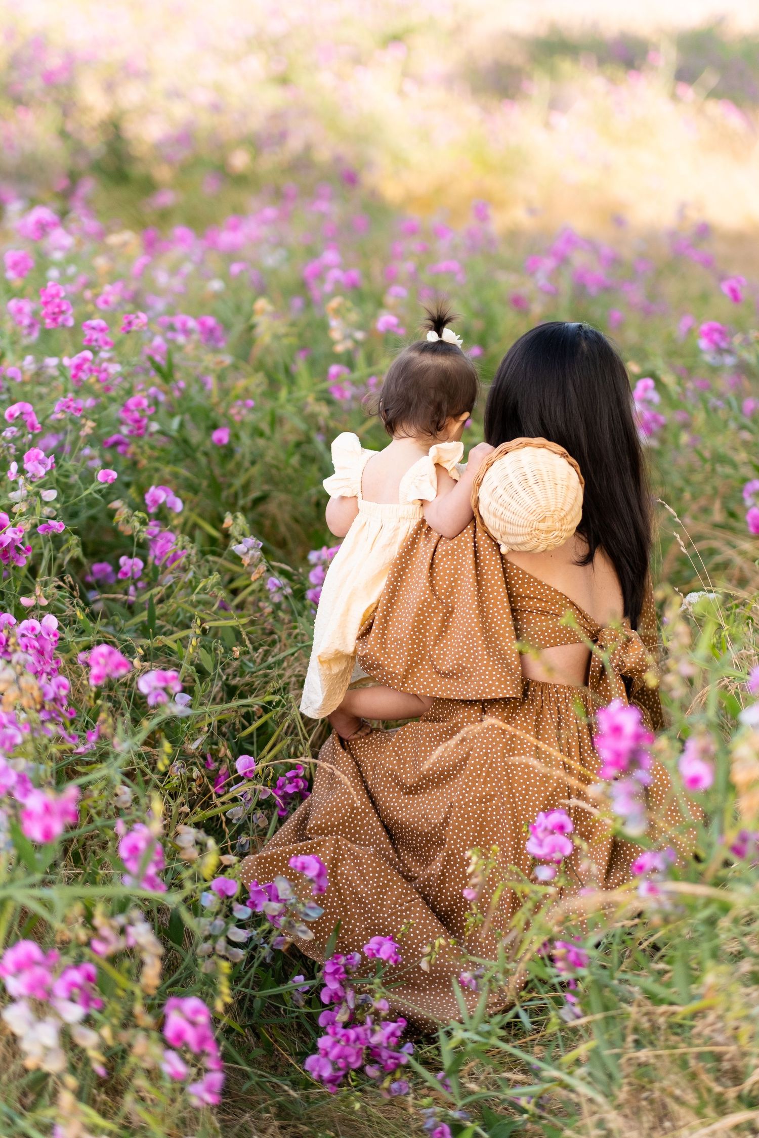 Two figures in polka dot dresses sit among vibrant purple wildflowers in a dreamy golden-hour meadow setting.