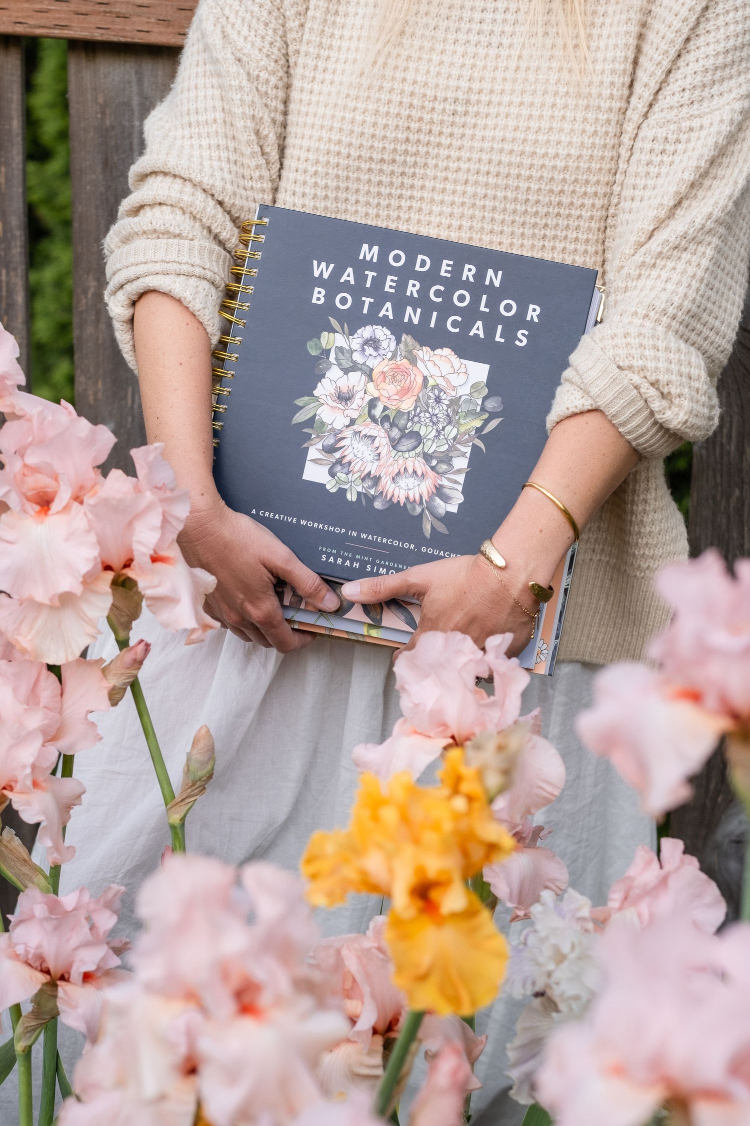 Woman holding Modern Watercolor Botanicals book by Sarah Simon surrounded by pink and yellow iris flowers.