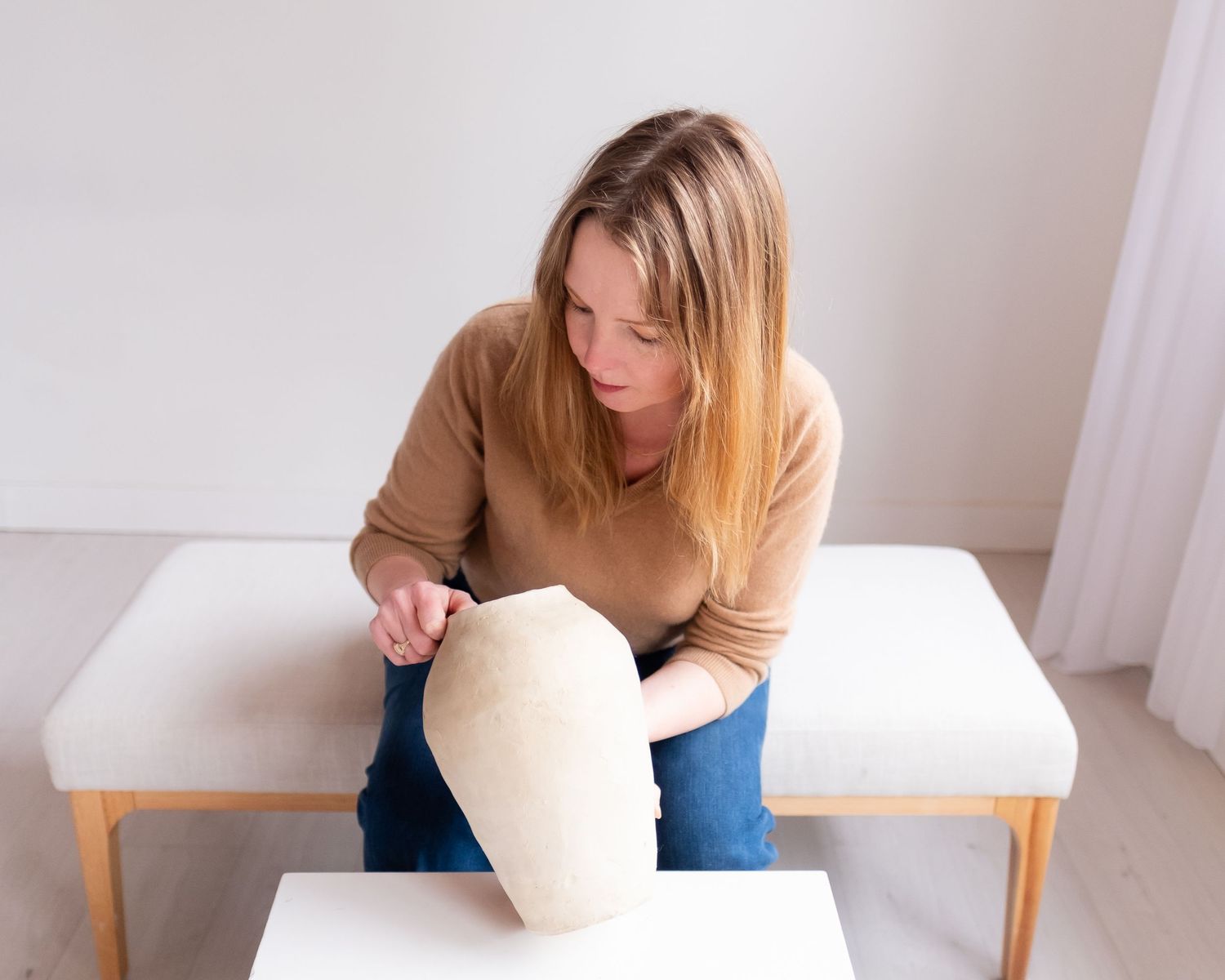 Someone in casual attire sits on a white bench examining a decorative white object in a minimalist interior setting.