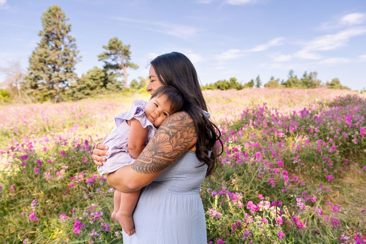 People embrace in a field of purple wildflowers on a sunny day with pine trees in the background.