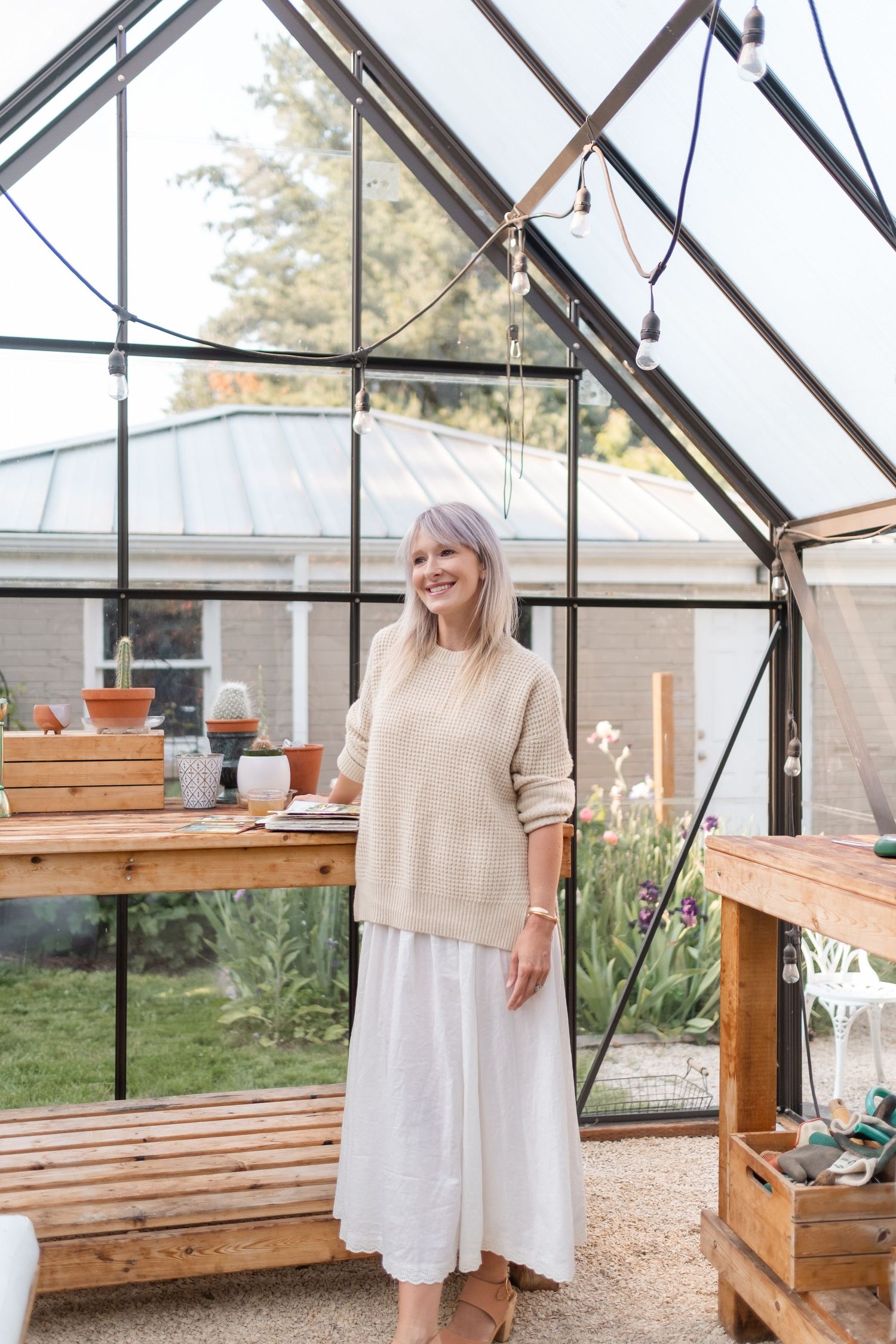 Woman in beige sweater and white skirt smiling inside a glass greenhouse with string lights and wooden potting benches.
