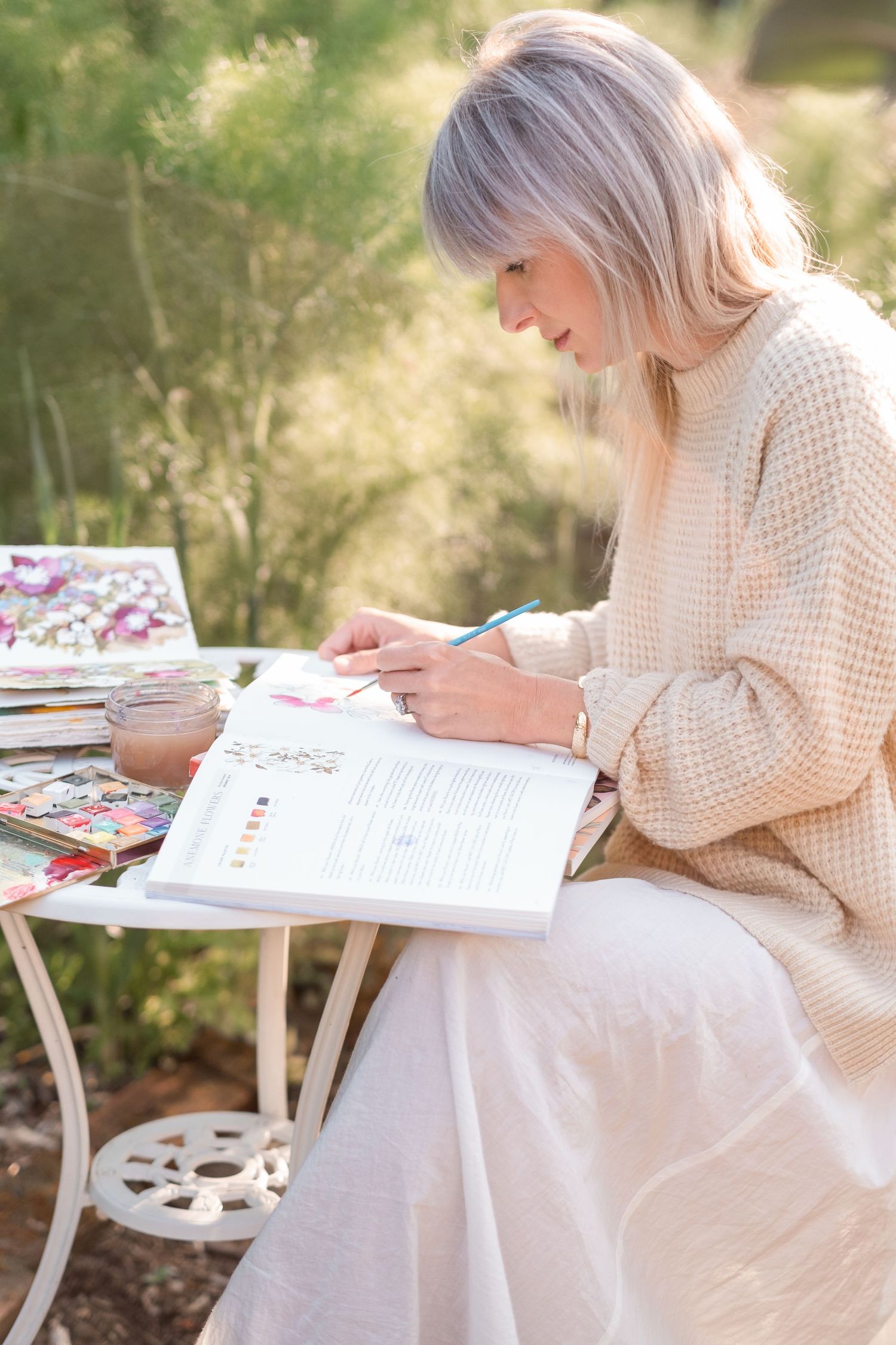 Woman in beige sweater writing in book at white garden table outdoors.