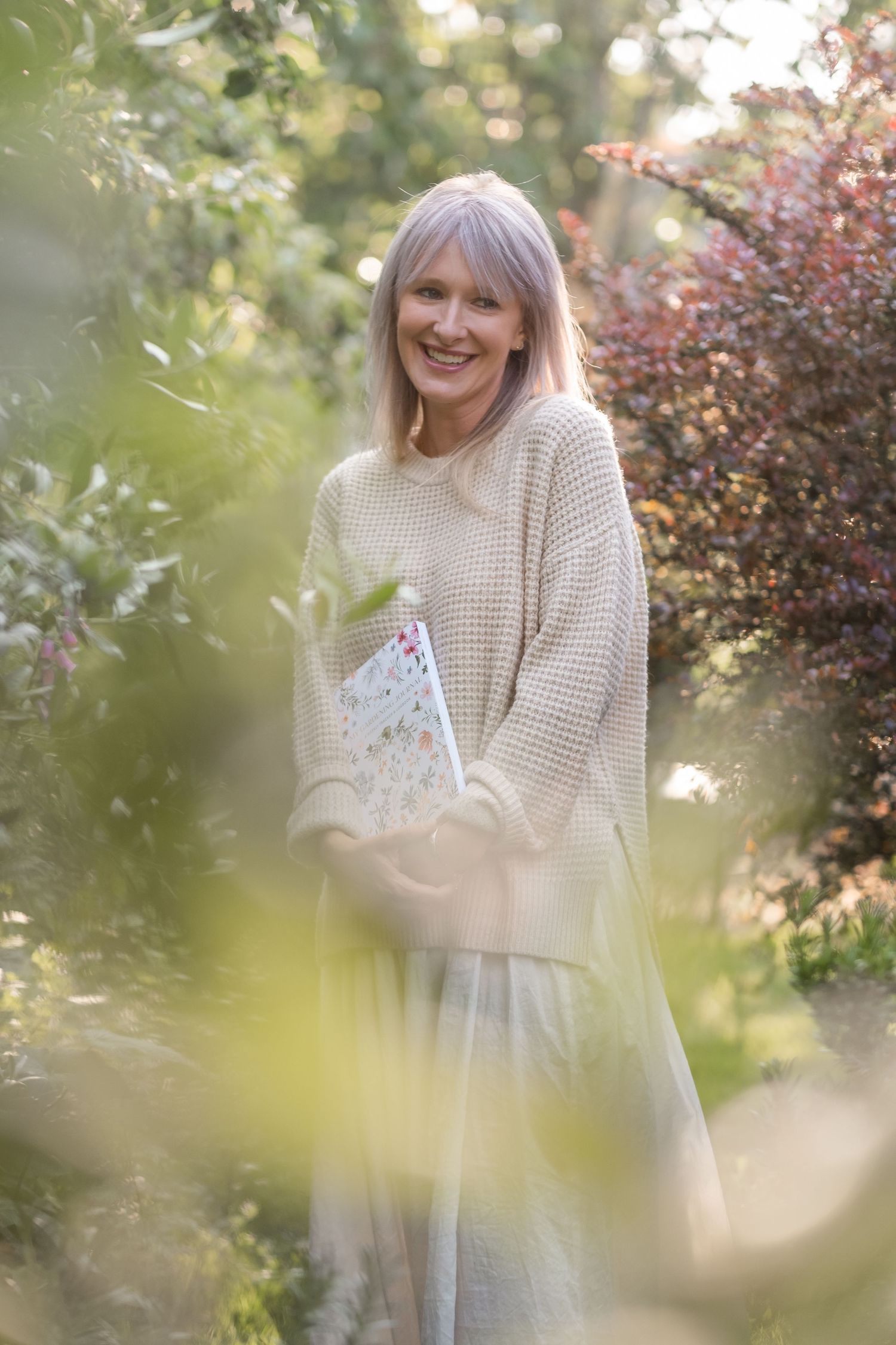 Smiling woman in beige sweater holding book while standing among lush green garden foliage.