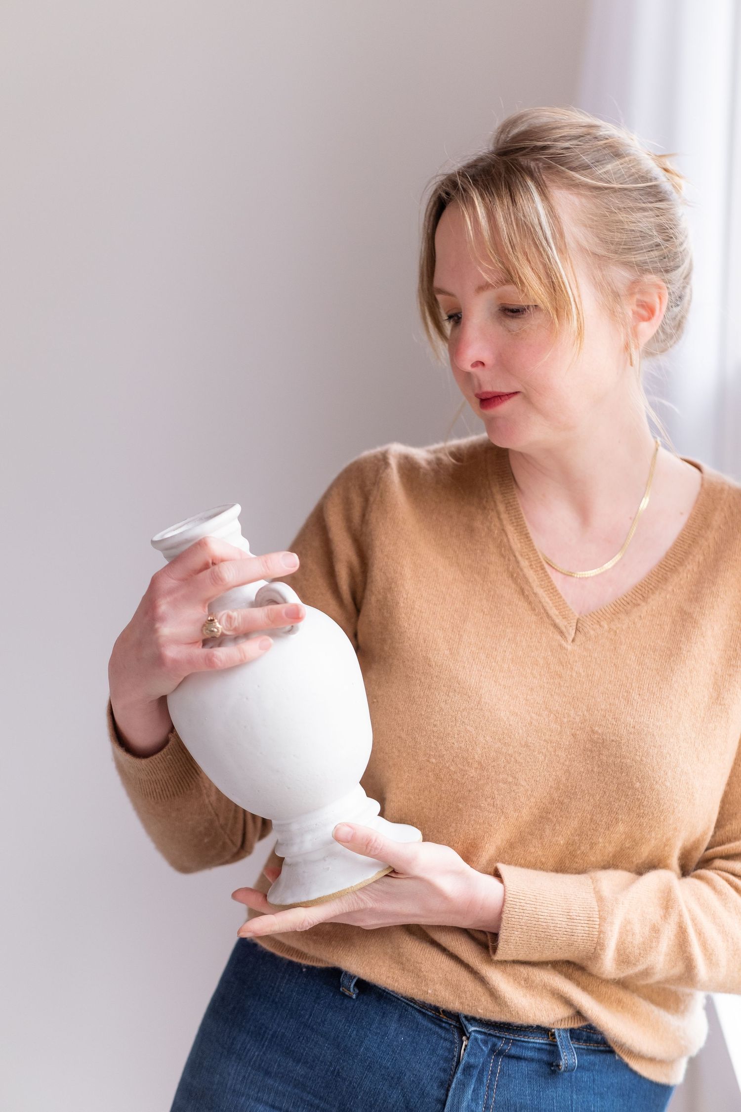 A ceramicist inspects a white pottery vessel while wearing a camel colored sweater in a bright studio.