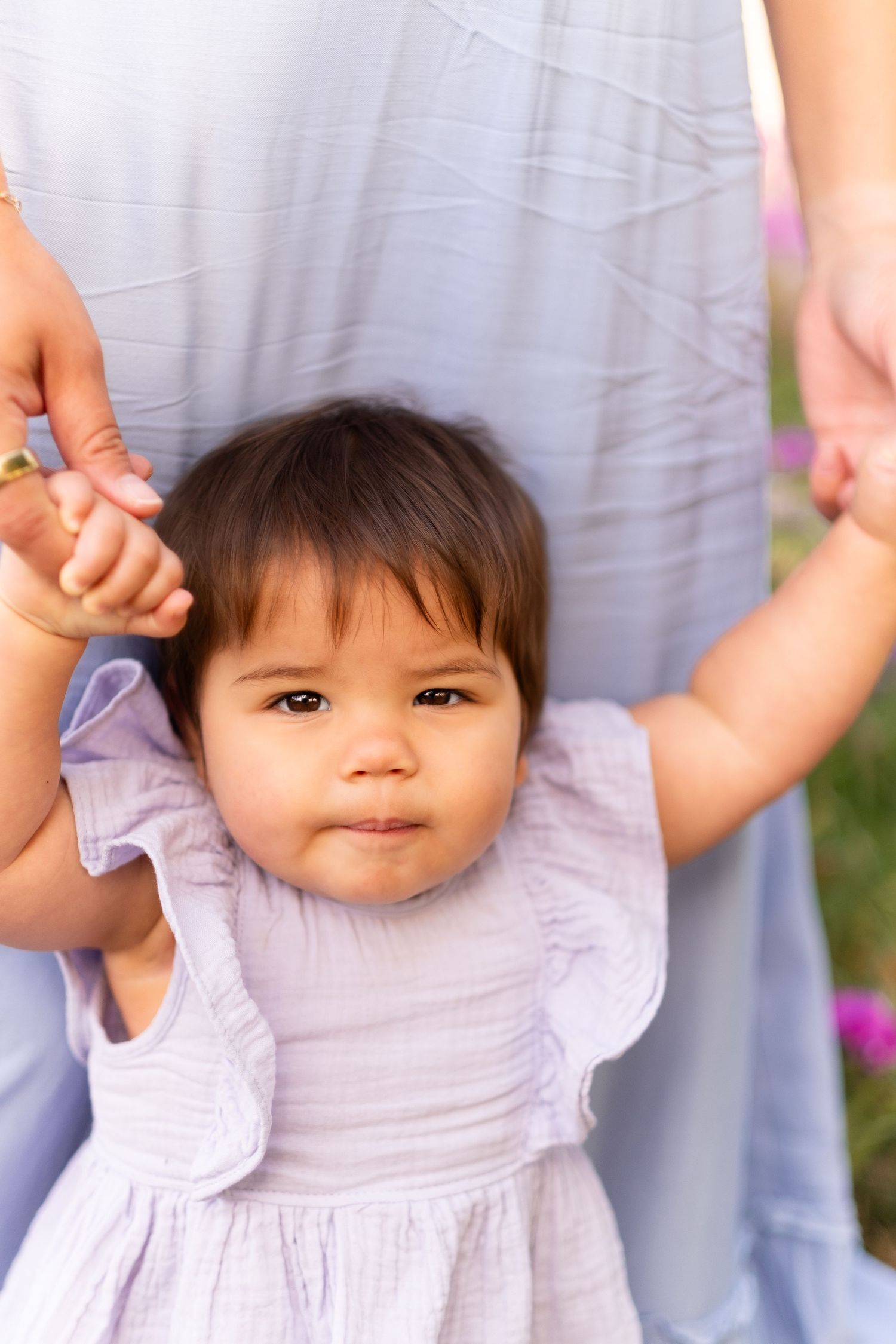 A small child in a light purple dress holds hands with adults while standing against a gray background.