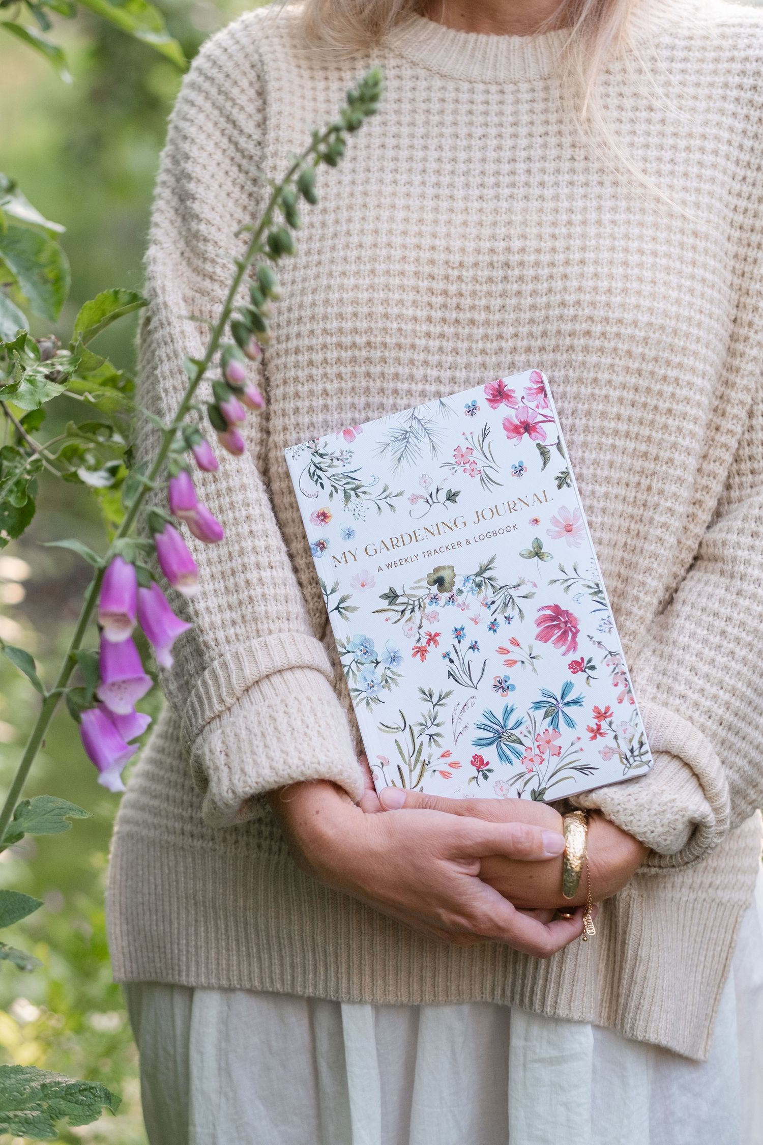 Woman in beige knit sweater holding a floral My Gardening Journal beside purple foxglove flowers outdoors.