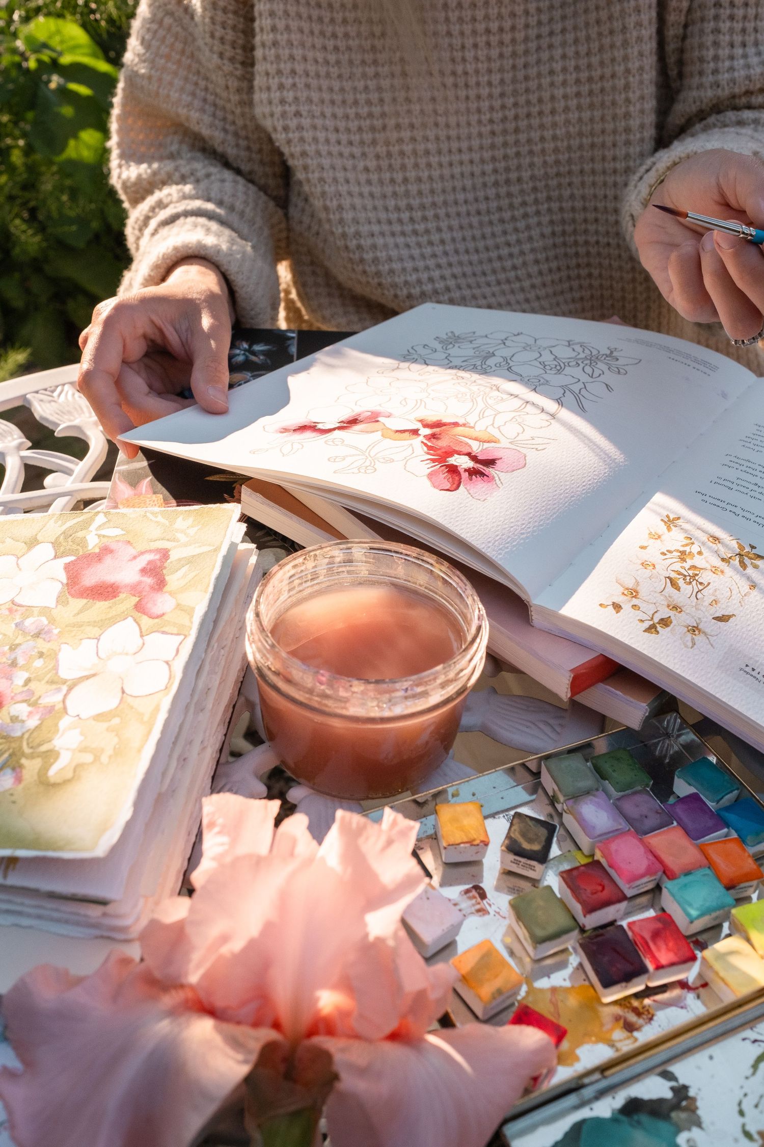 Close-up of hands browsing floral illustration book with watercolor paints and candle on garden table.