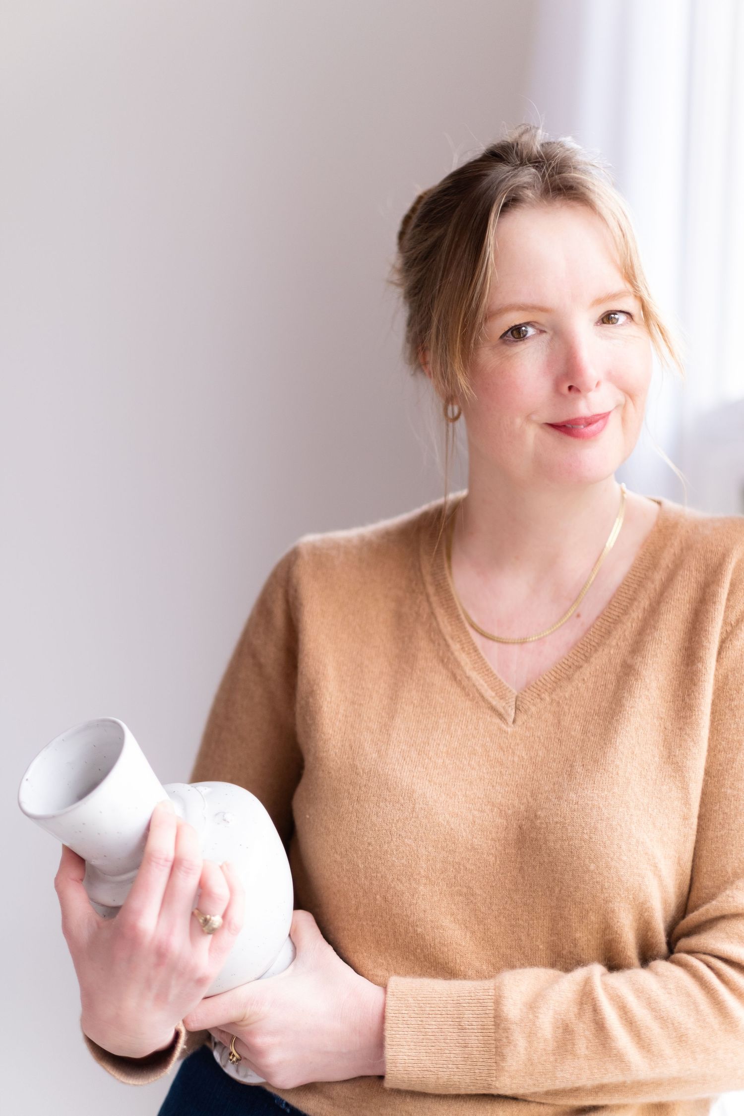 Person in tan sweater holding white ceramic vessel against bright window.
