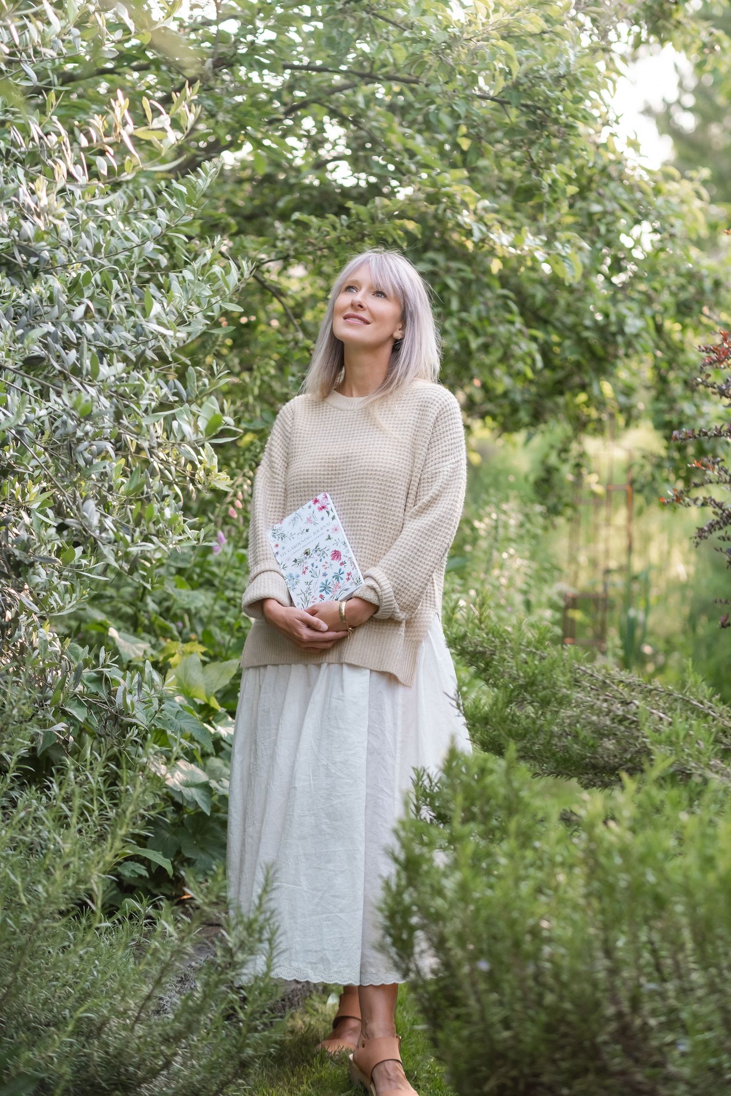 Blonde woman in beige sweater holding floral book looking up in green garden.