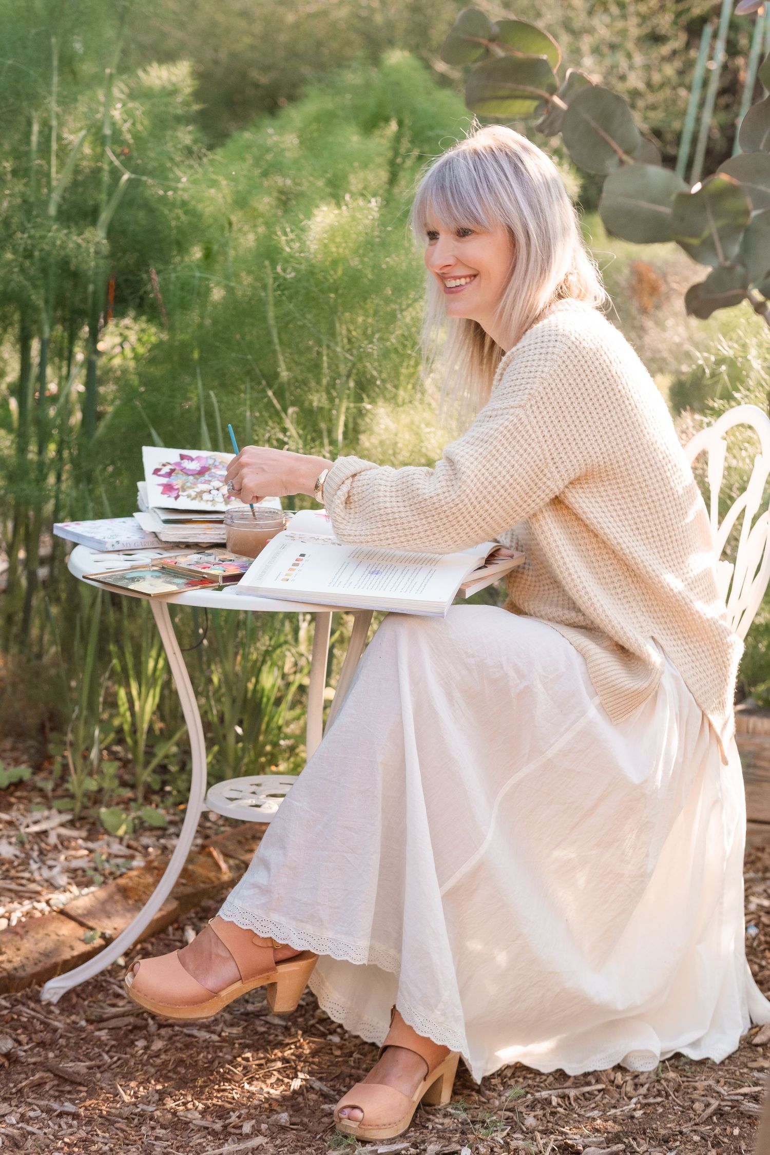 Woman in beige sweater sitting at white garden table with watercolor supplies, smiling outdoors.