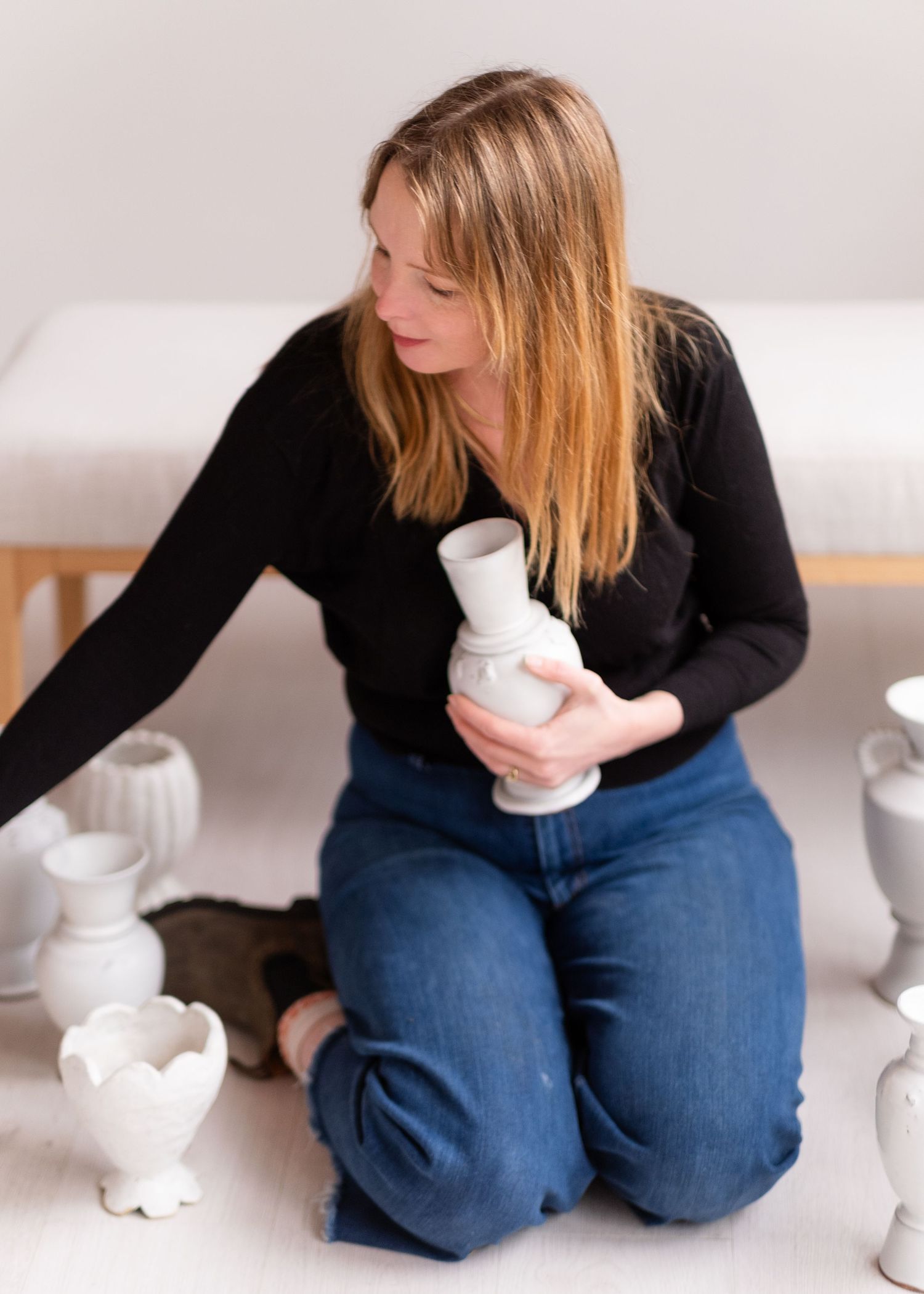 Artist examines white ceramic pottery while sitting on floor in black sweater and blue jeans.
