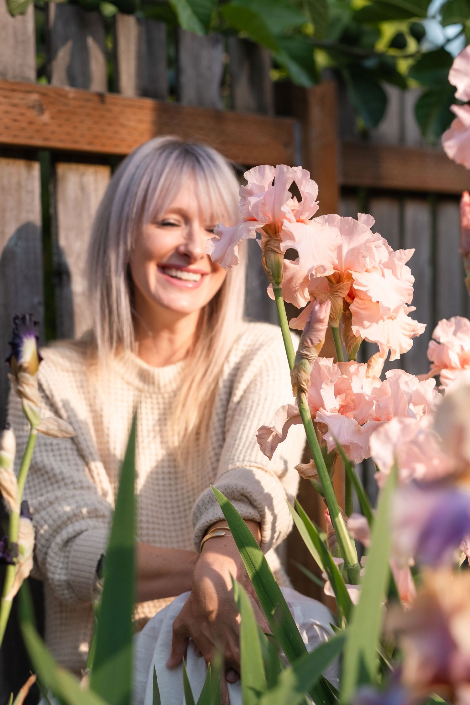Smiling blonde woman in cream sweater admiring pink iris flowers in sunny garden.