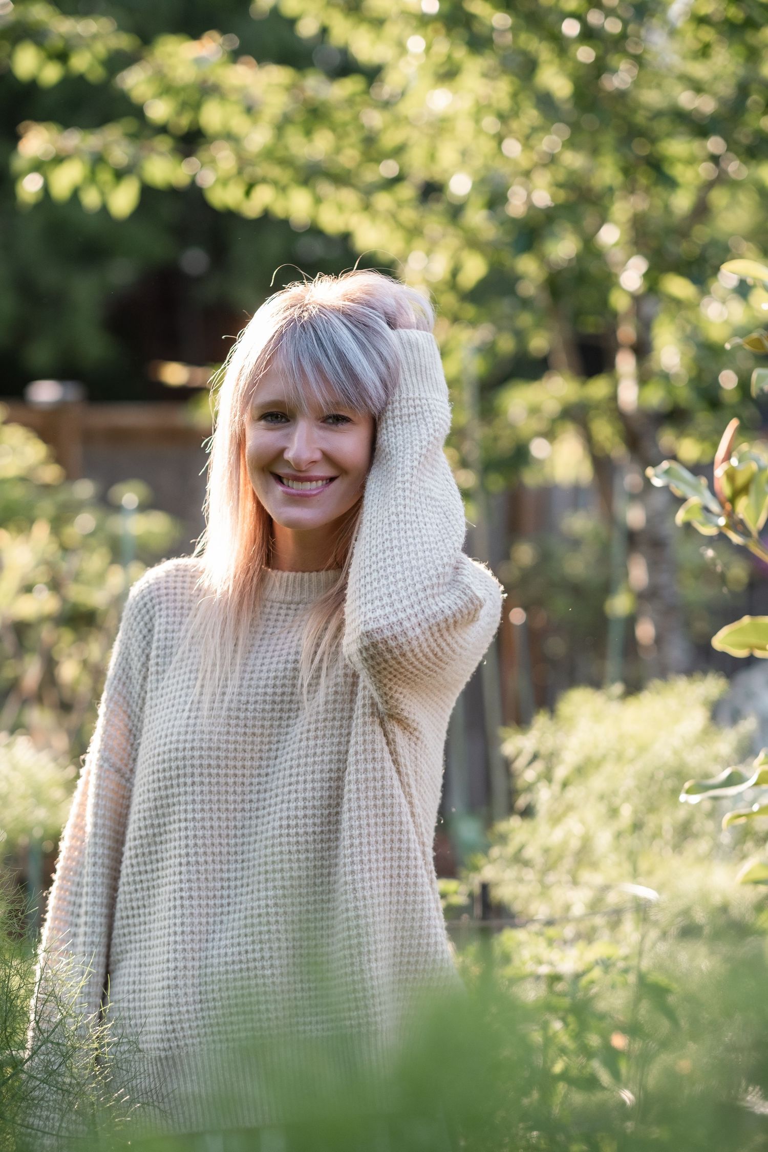 Happy blonde woman in cream knit sweater standing in lush green garden with hand behind head.