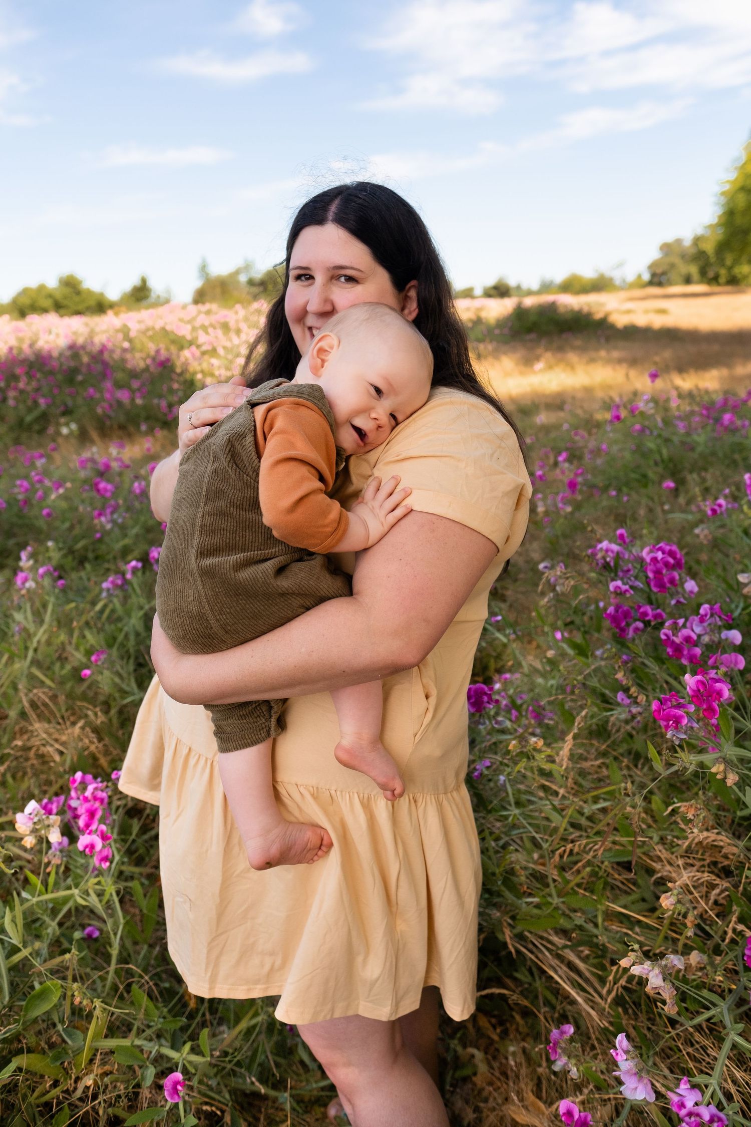 A woman holds a sleeping baby in a yellow dress while standing in a field of vibrant purple wildflowers.