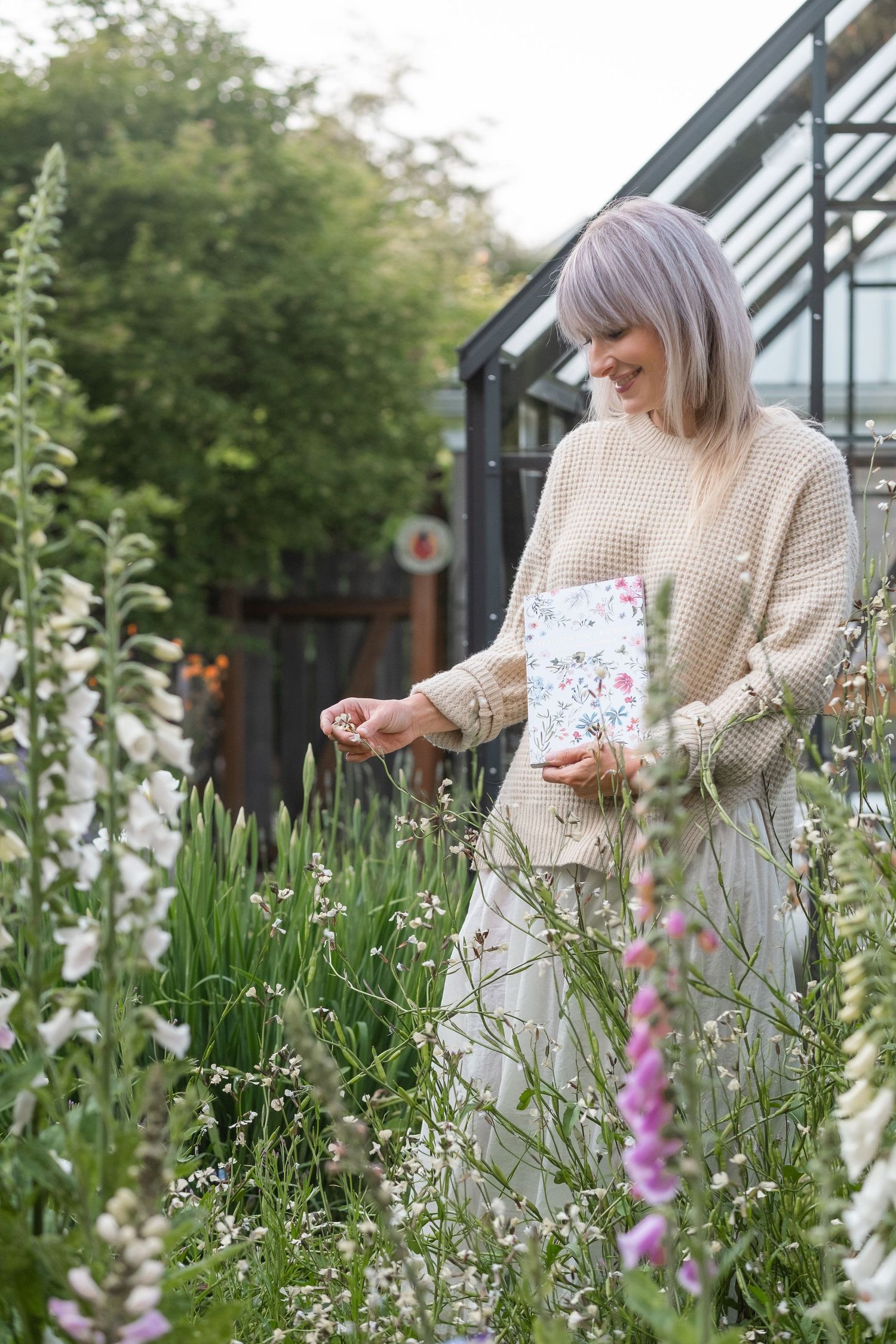 Smiling woman in beige sweater holding a floral notebook while walking through a garden of wildflowers.