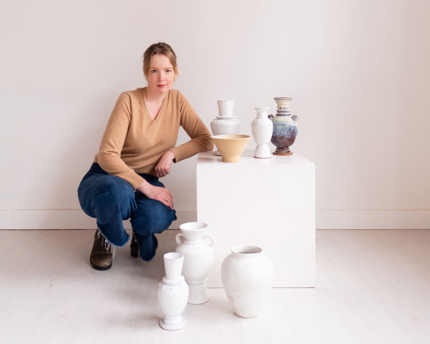 Artist poses with handmade ceramic vessels and sculptures displayed on a white pedestal against neutral wall.