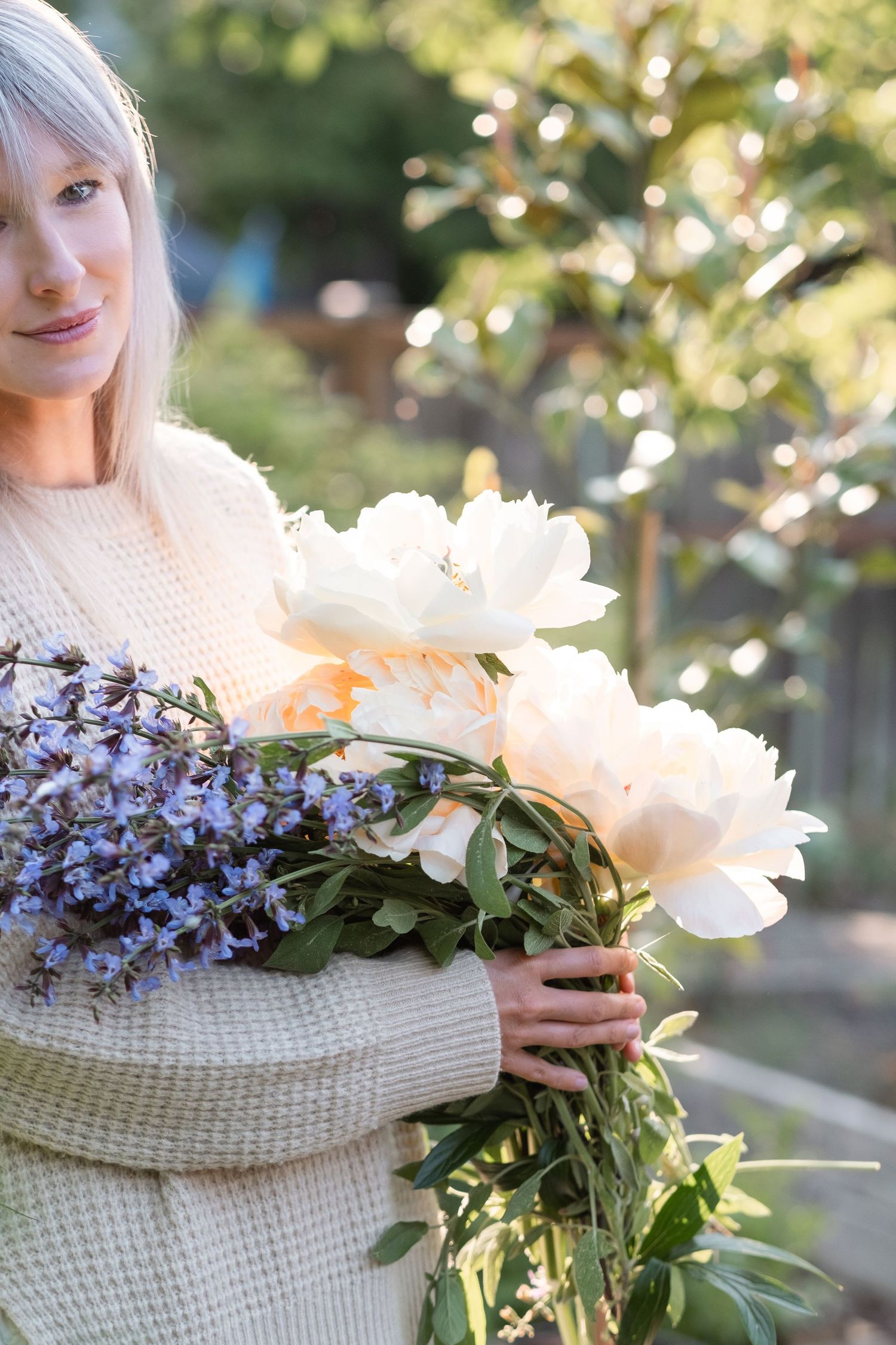 Woman in cream sweater holding large bouquet of white peonies and purple flowers in sunny garden.
