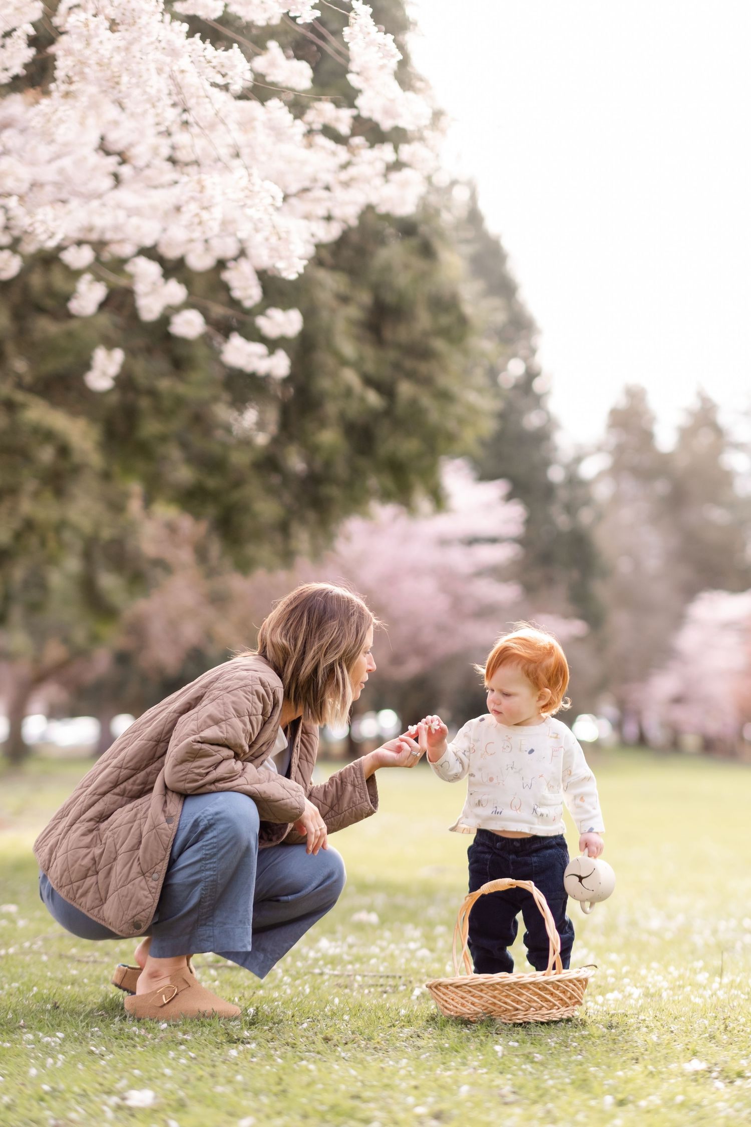 Mother and toddler sharing a tender moment in a cherry blossom park, with a wicker basket on the grass.