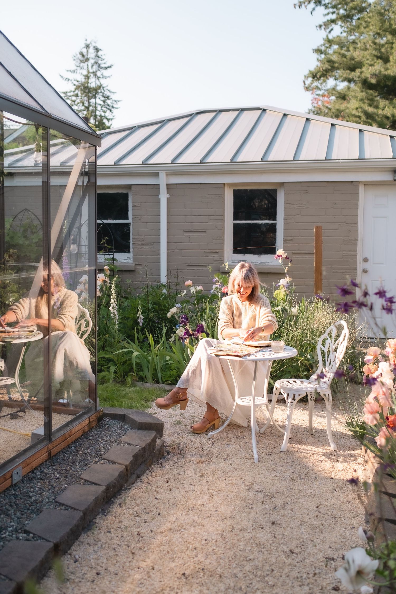 Woman sitting at white garden table reading near greenhouse and flower garden.