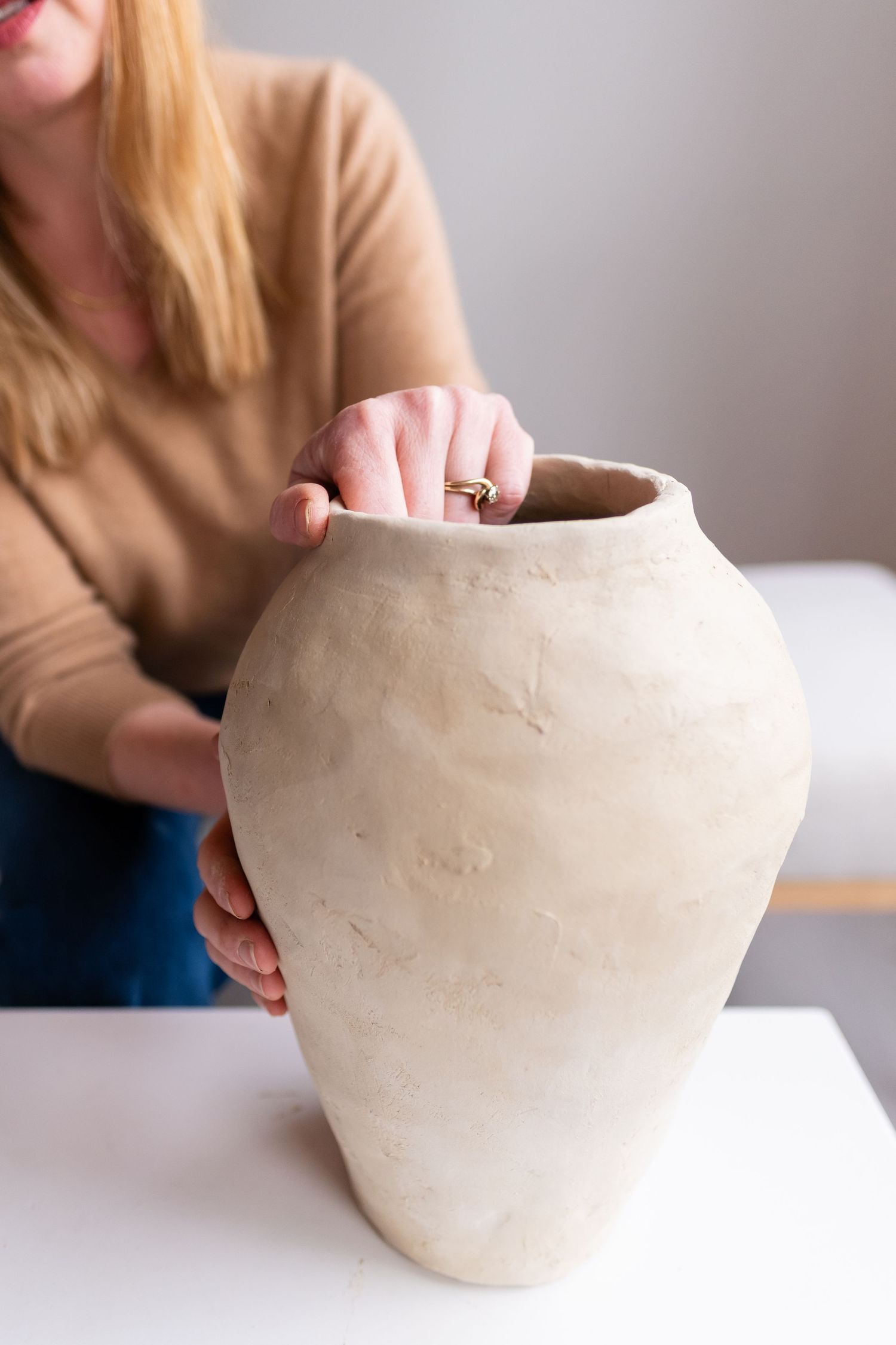 Hands holding and examining a raw clay pottery vase on white table.