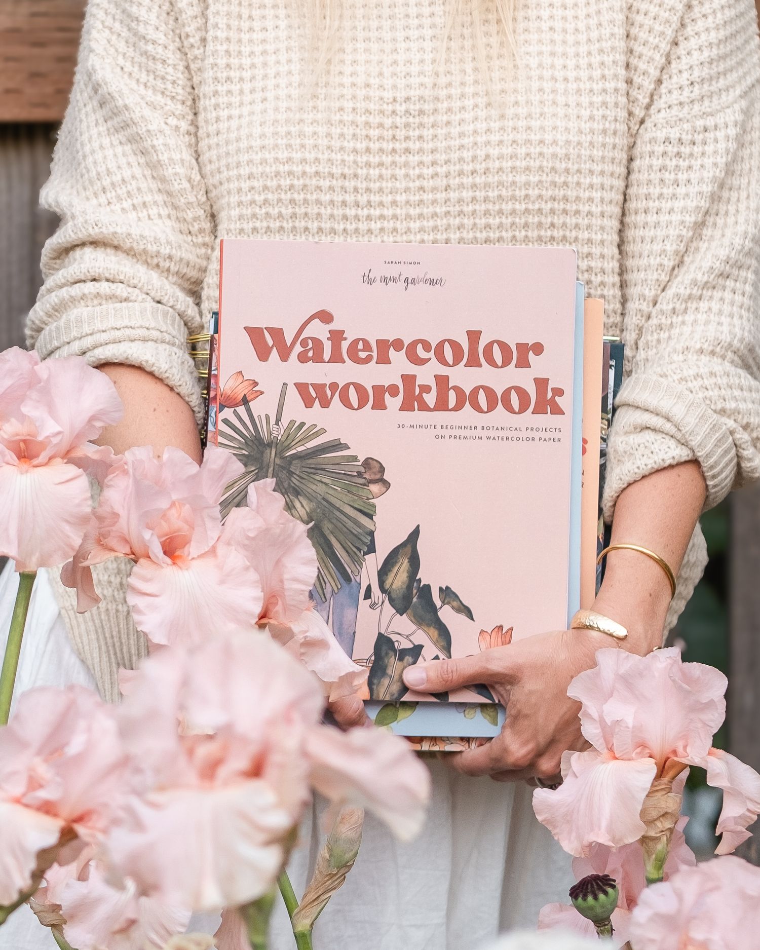 Person holding a pink Watercolor Workbook by The Rooftop Muse surrounded by soft pink iris flowers in bloom.