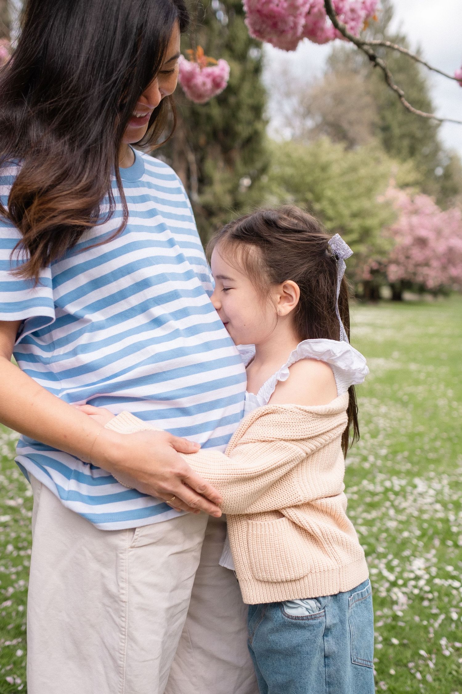 A touching moment between a pregnant woman in a striped shirt and a young child embracing under blooming cherry trees.