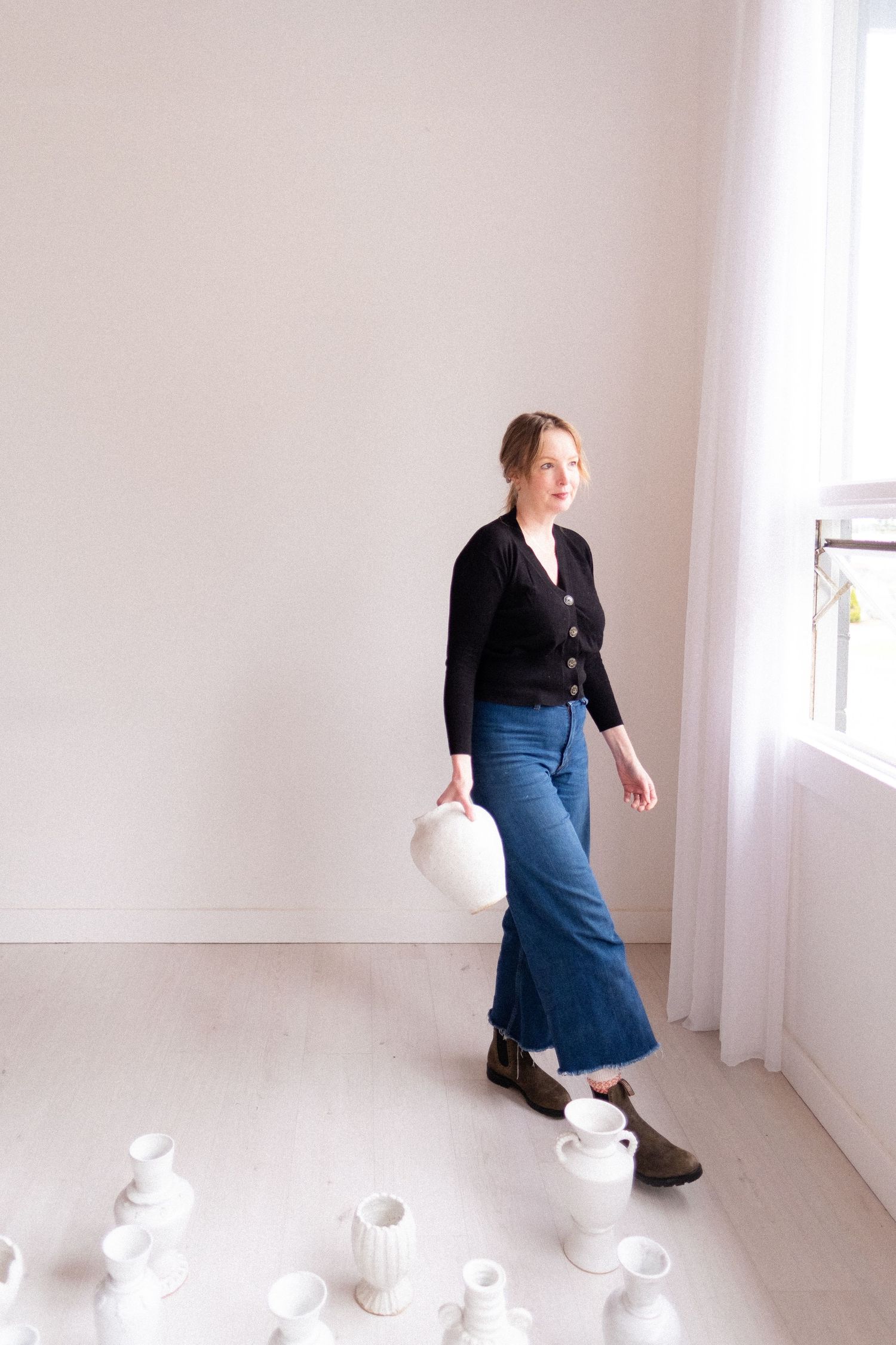 Person in black cardigan and wide-leg jeans stands in minimalist room with white ceramic objects on floor.