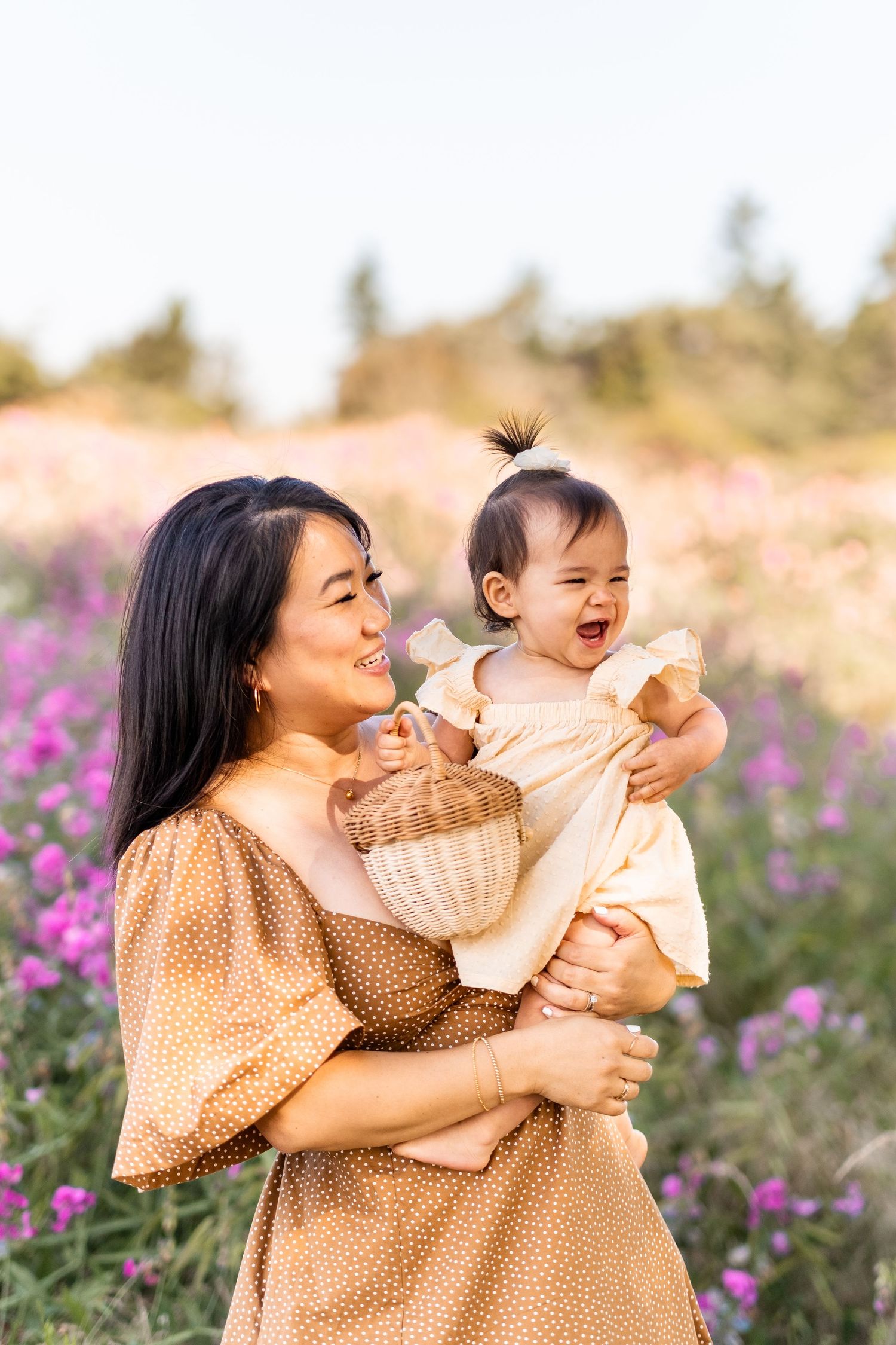 A joyful moment shared between a woman in tan dress and laughing baby in a field of purple flowers.