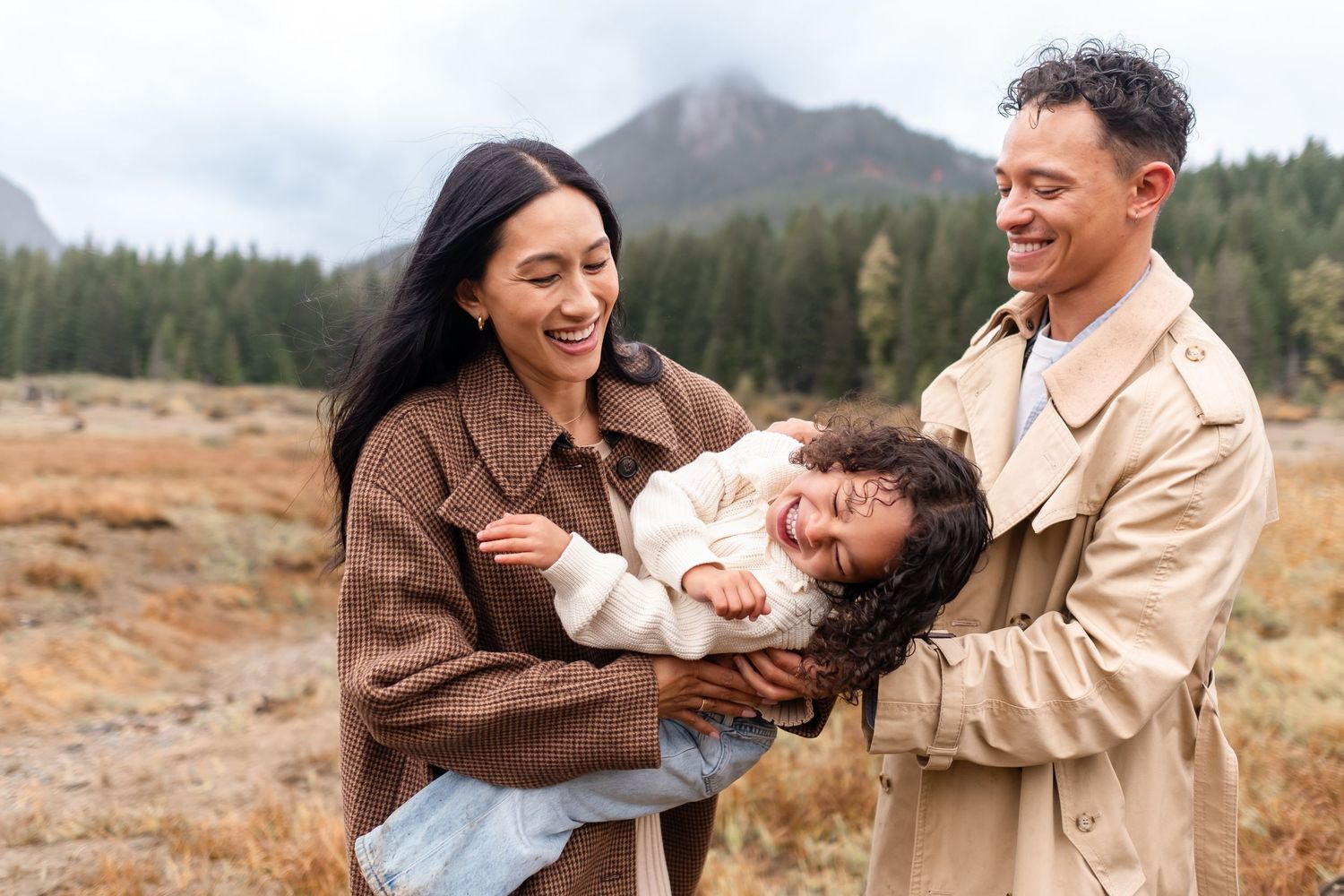 Happy family with parents laughing and holding their young child outdoors in a scenic mountain meadow setting.