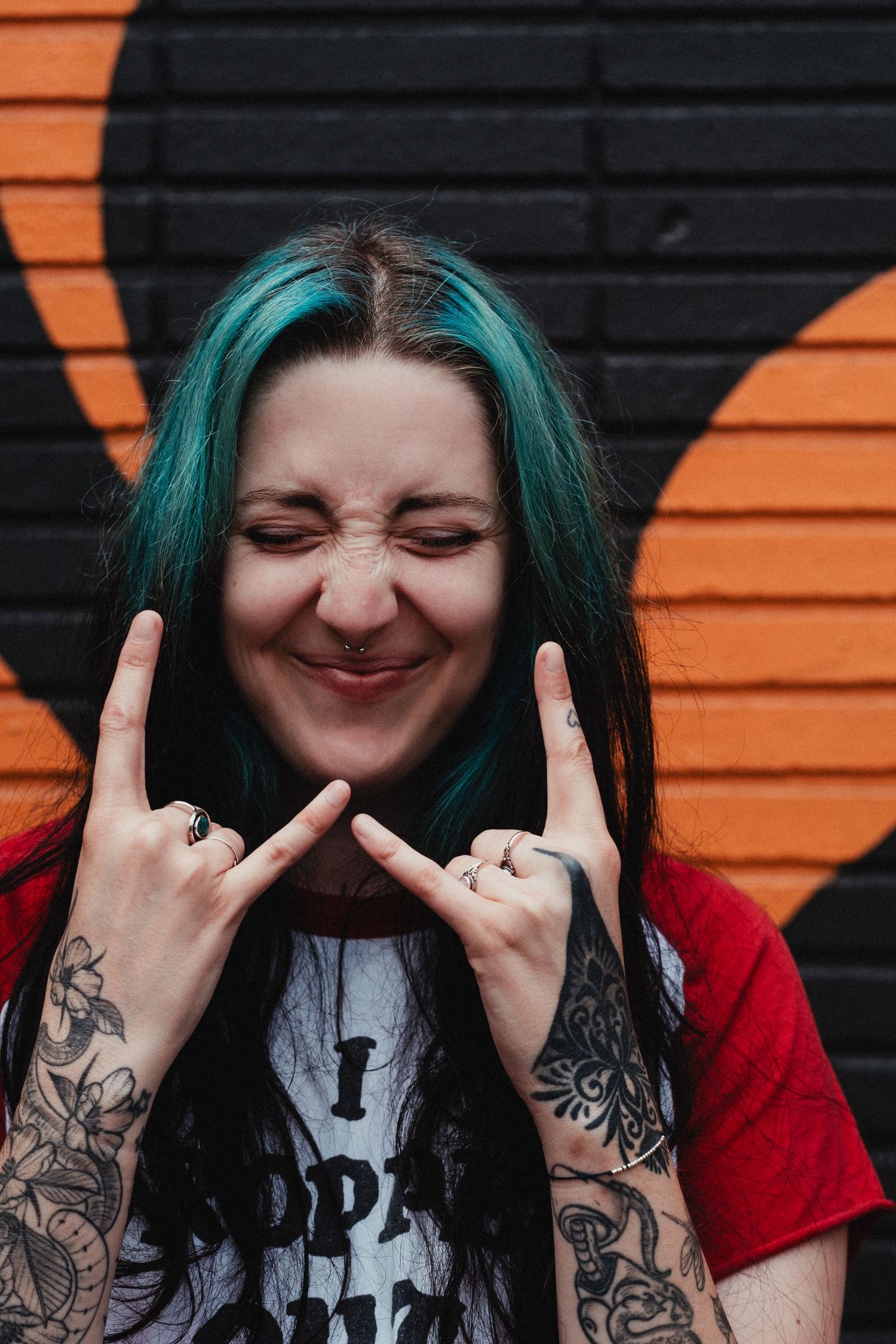 Tattooed person with teal hair and red shirt making rock and roll hand gestures against orange and black wall.